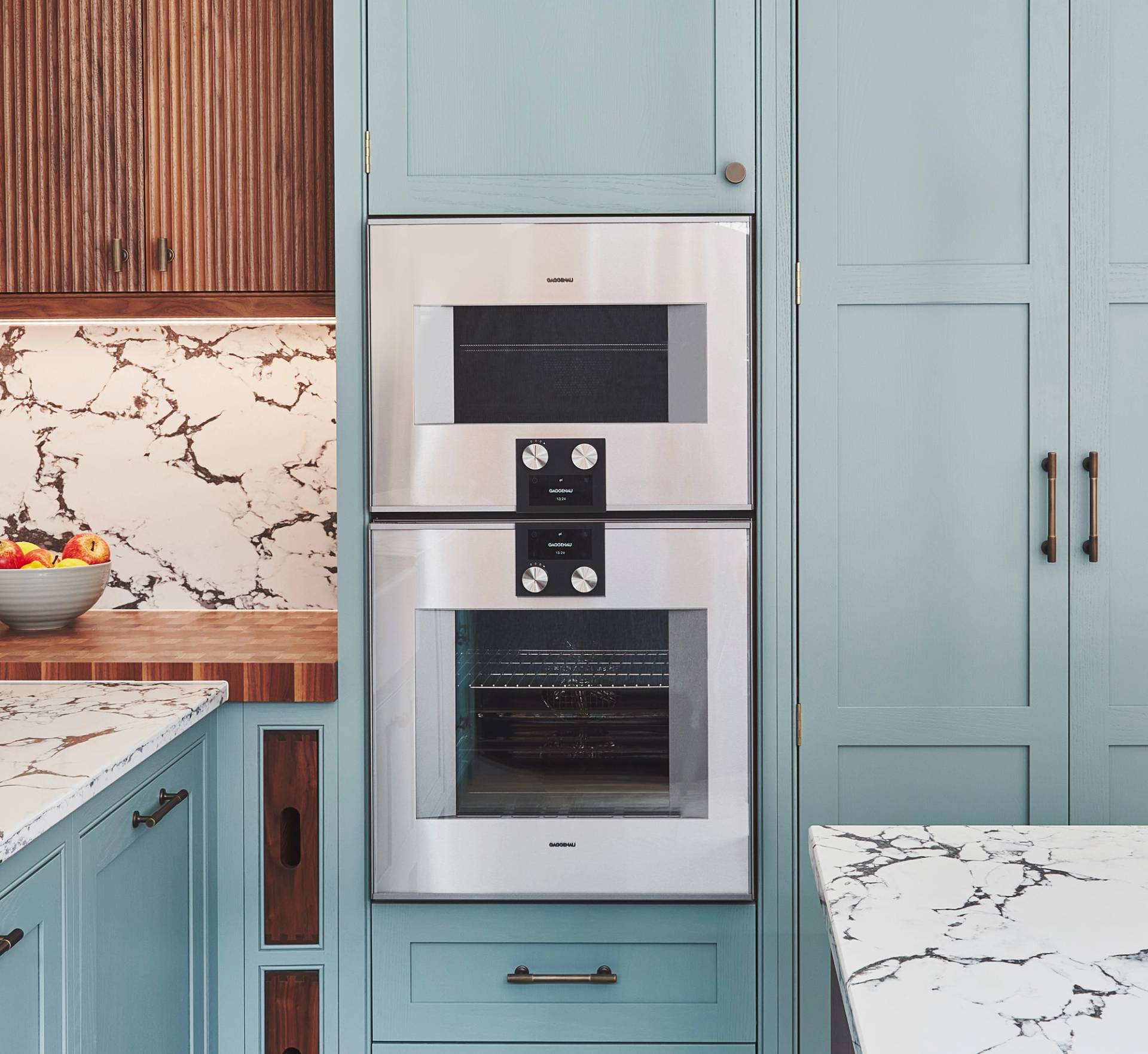 Modern kitchen with built-in double oven set in light blue cabinetry. Surrounding countertops and backsplash feature a white and gray marble pattern. A bowl filled with fruit sits on the left countertop.