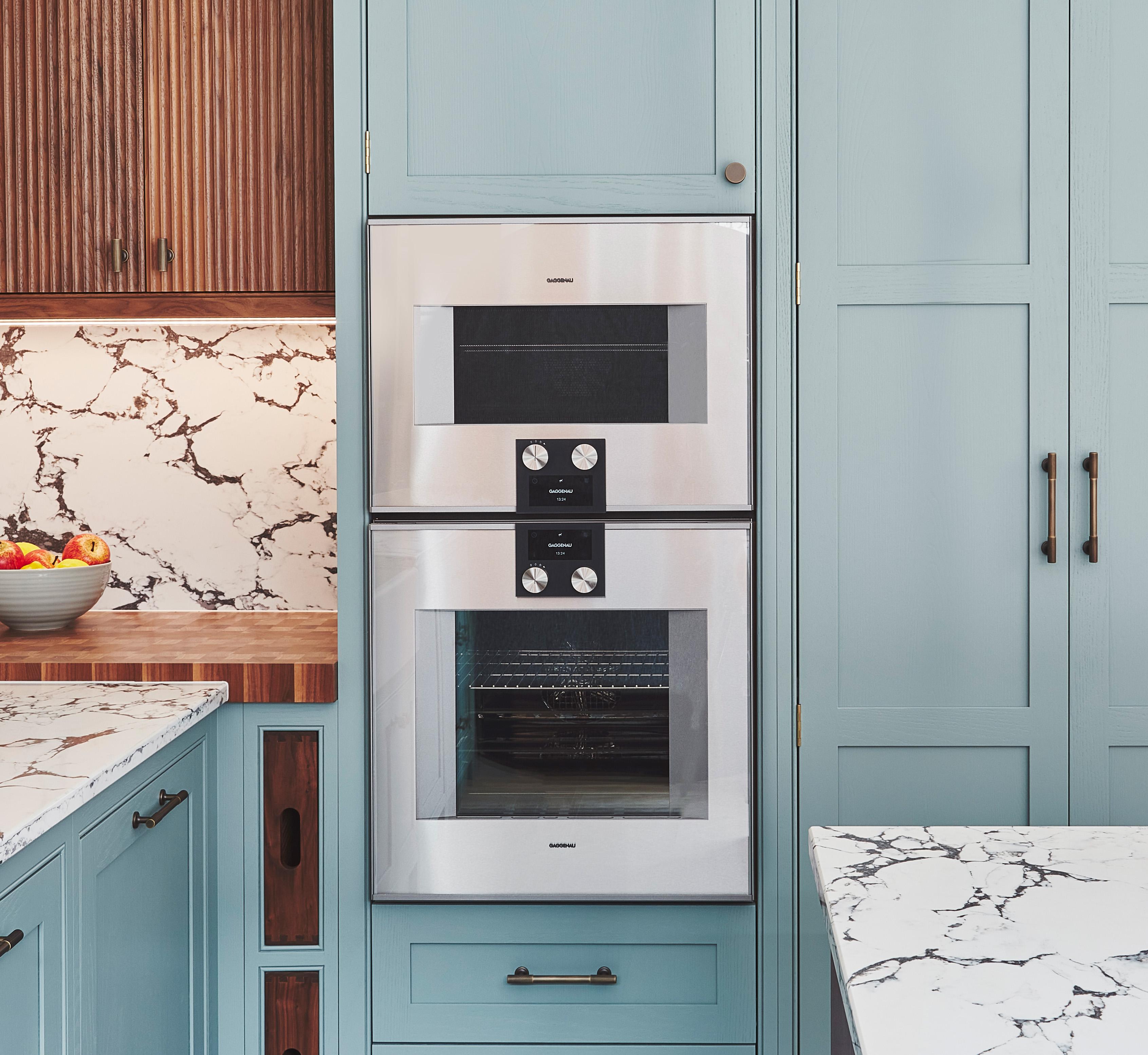 Modern kitchen with built-in double oven set in light blue cabinetry. Surrounding countertops and backsplash feature a white and gray marble pattern. A bowl filled with fruit sits on the left countertop.