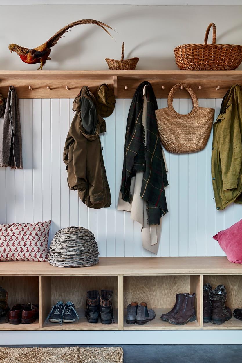 A boot room with a wooden bench, various boots and shoes in cubbies, coats and scarves hanging on hooks, baskets on a shelf, and a pheasant display on top. Cushions sit on the bench.