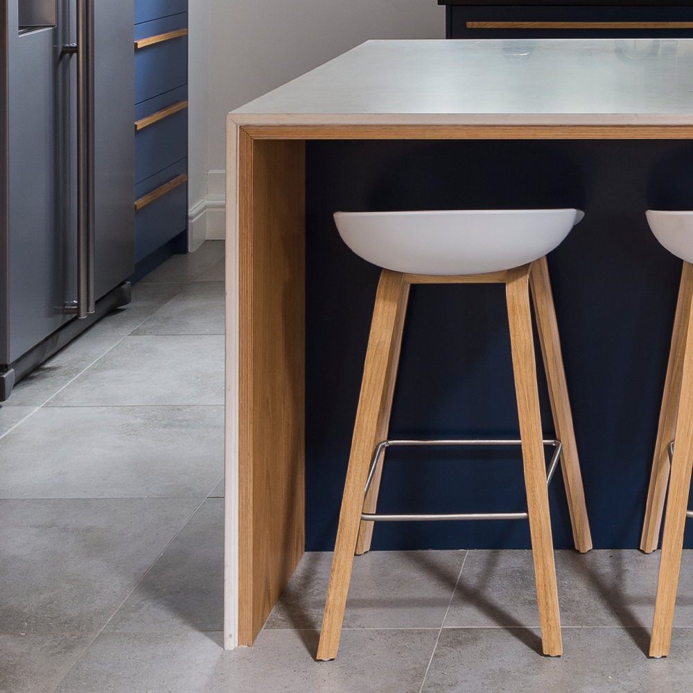 Modern kitchen with a wooden and navy blue island, featuring two white bowl-shaped bar stools with wooden legs on a gray tiled floor. A stainless steel refrigerator is partially visible on the left.