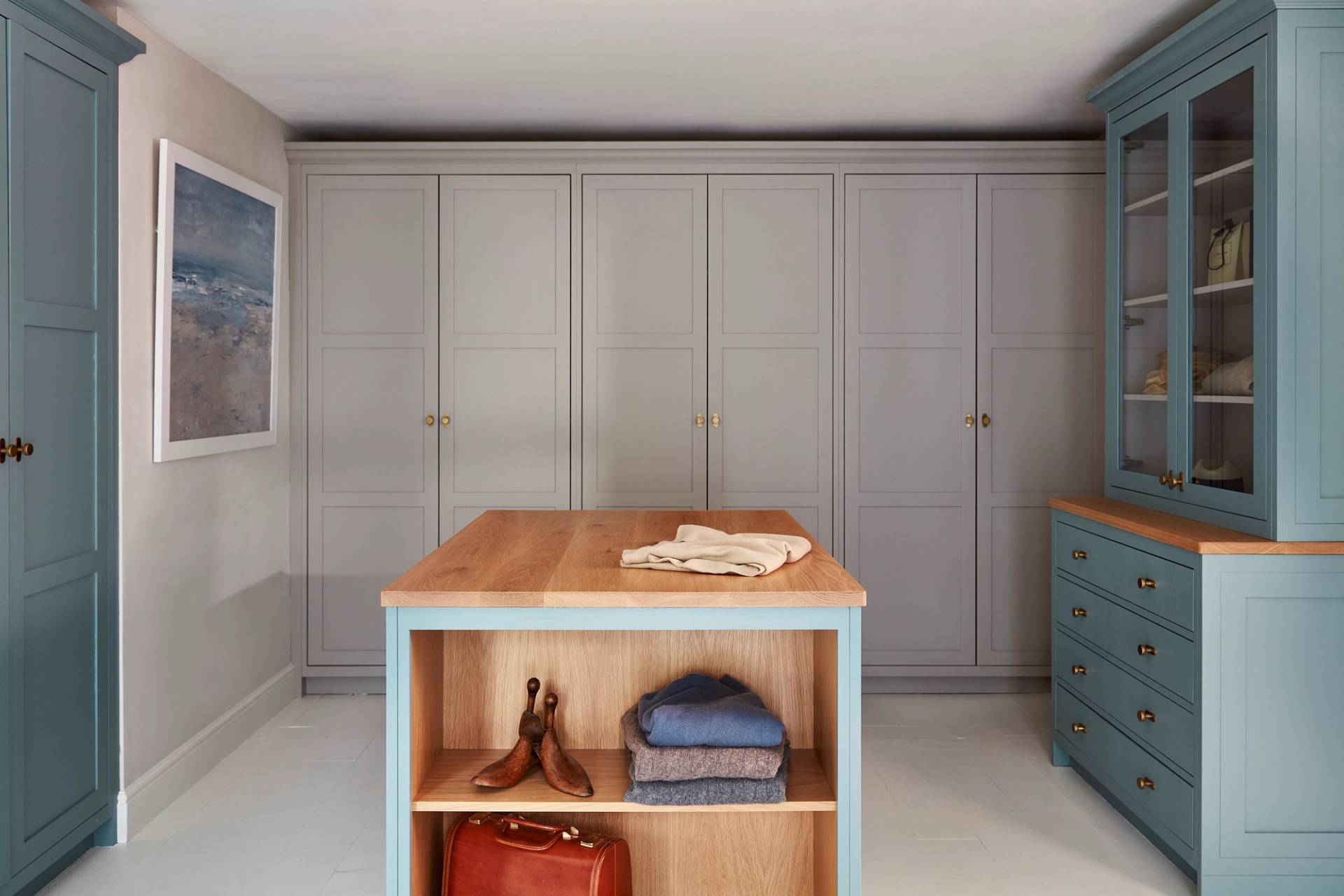 A tidy, modern walk-in closet with light gray cabinets, a wooden-topped island with folded clothes, shoes, and a brown bag on its shelves. Blue cabinetry and a framed wall art complete the room.