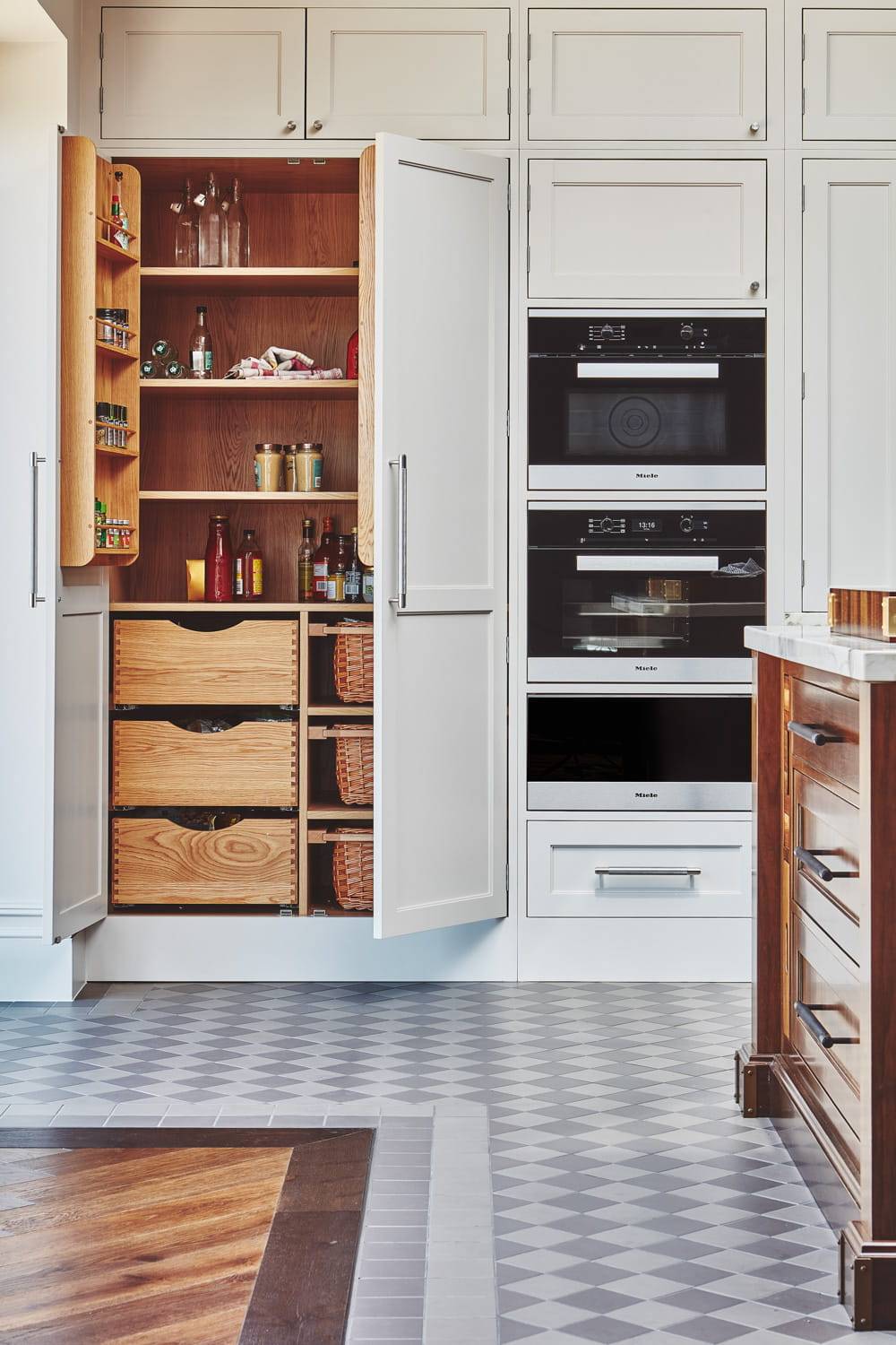 A kitchen with a tall white cabinet door open, revealing shelves with various jars, bottles, and woven baskets. Next to it are built-in stainless steel ovens. The floor features a gray and white checkered pattern.