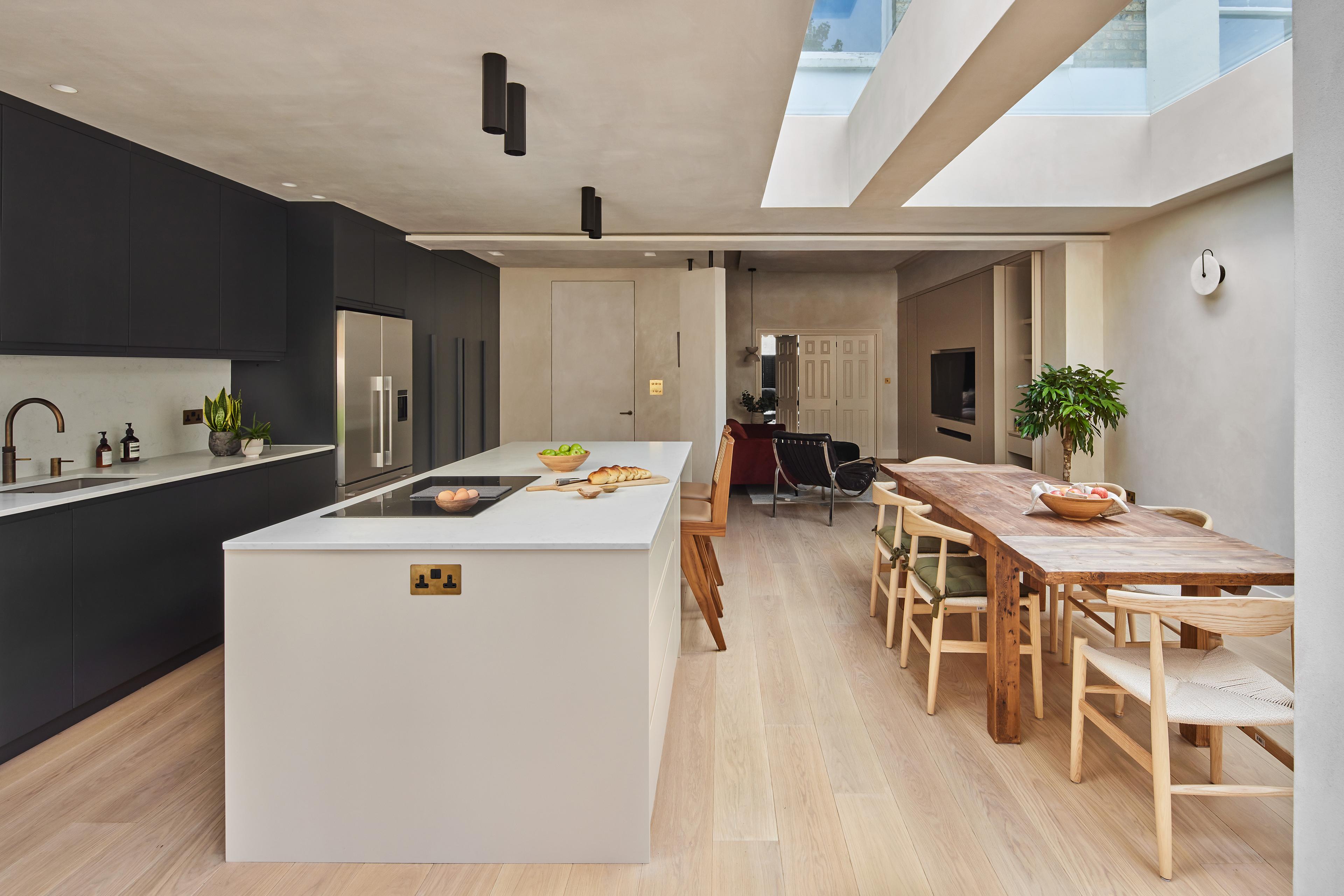 Modern open-plan kitchen and dining area with a large island, black cabinets, wooden dining table with chairs, light wooden floor, potted plants, and skylights letting in natural light.