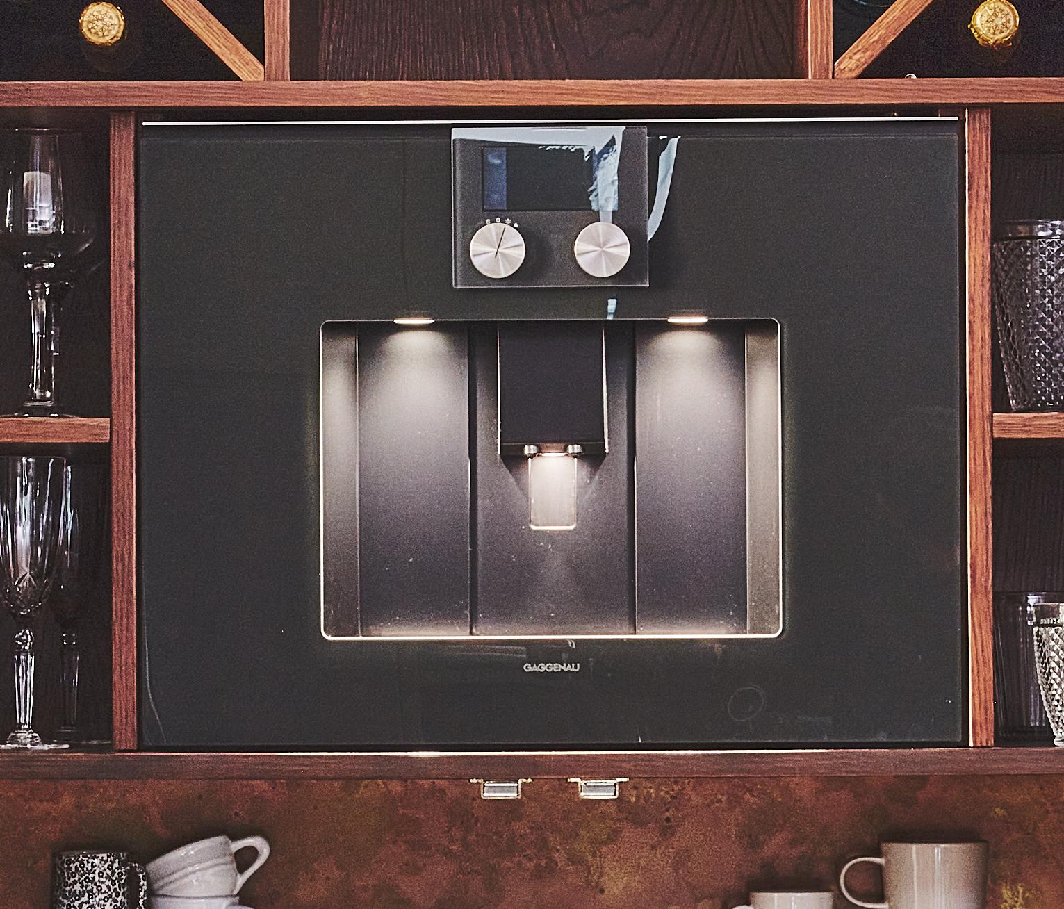 Built-in espresso machine with a sleek black front, two control knobs, and an illuminated dispensing area. Surrounded by wooden shelves holding glasses, mugs, and other barware.