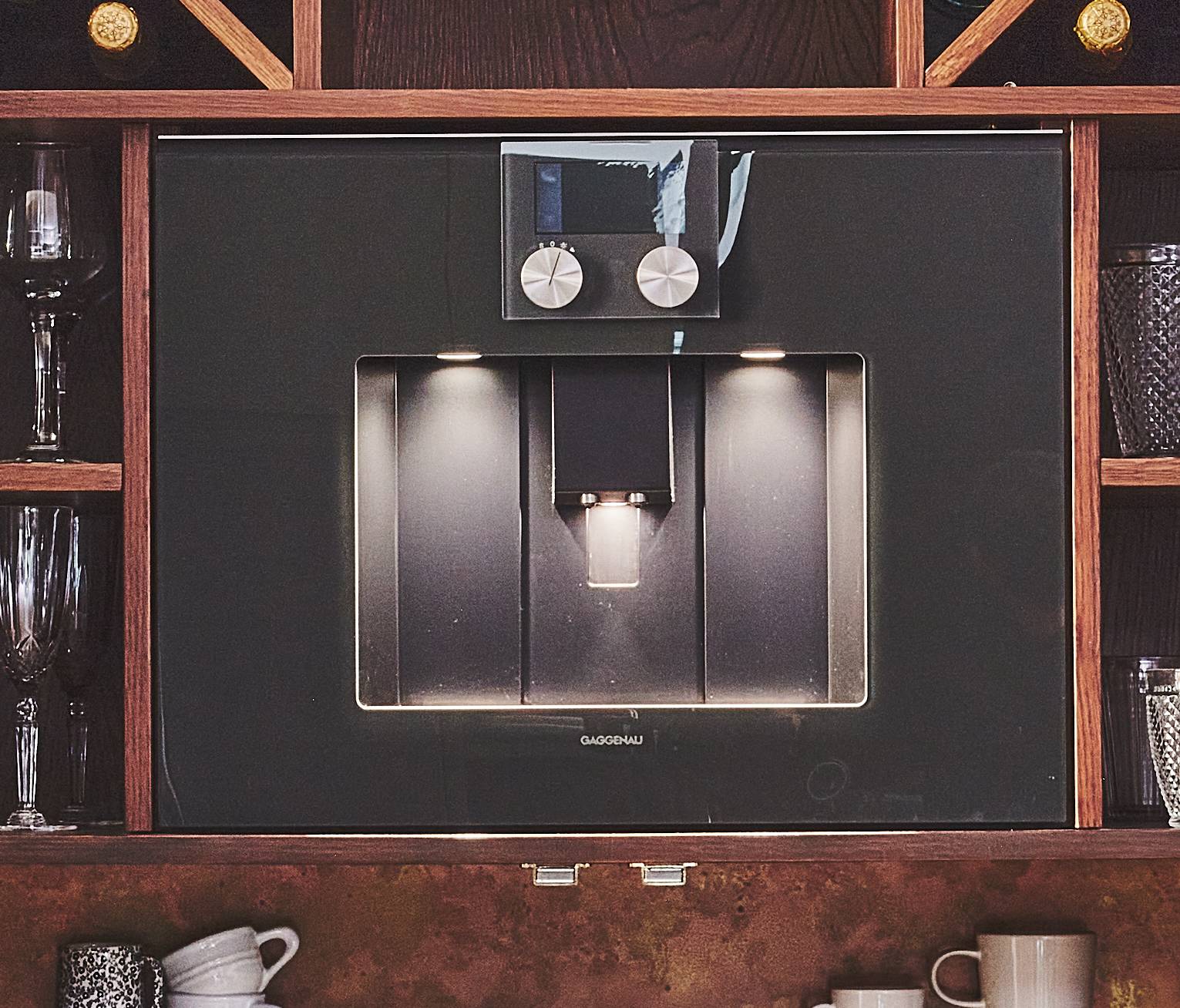 Built-in espresso machine with a sleek black front, two control knobs, and an illuminated dispensing area. Surrounded by wooden shelves holding glasses, mugs, and other barware.