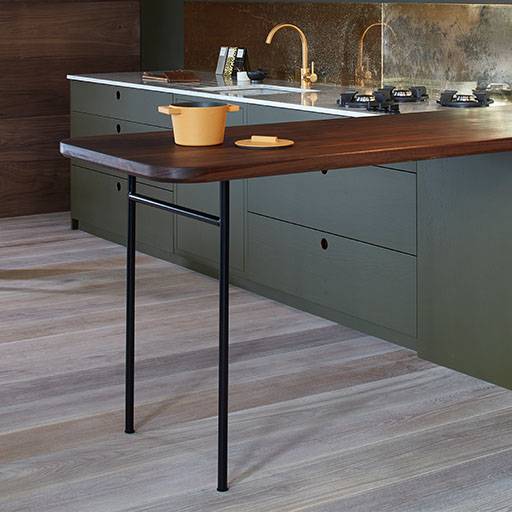 A modern kitchen with olive green cabinets, a wooden countertop extending as a breakfast bar with black metal legs, and a yellow pot and wooden spatula on top. The floor is light wood, and the backsplash is metallic.