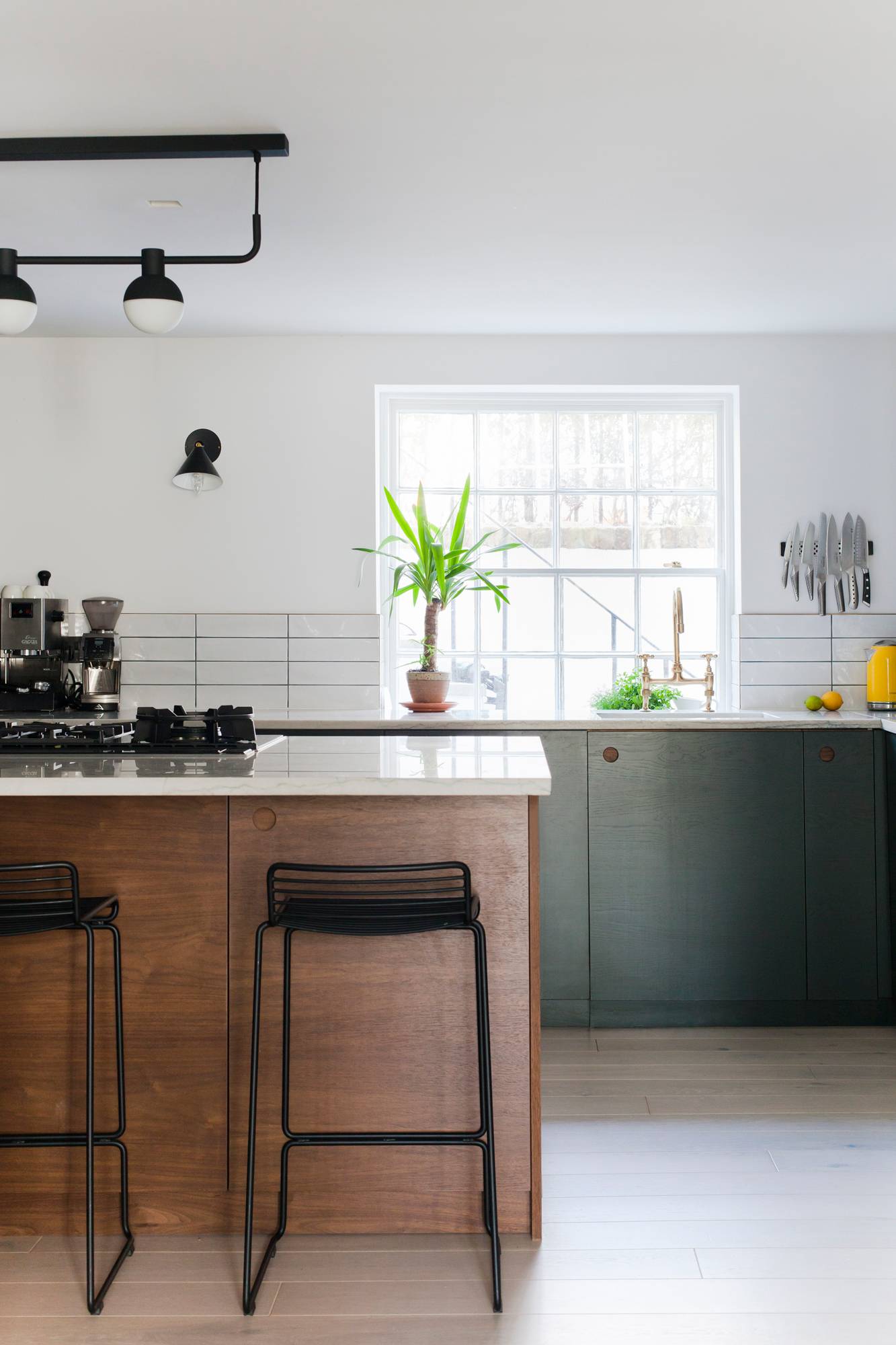 A modern kitchen with a central island featuring two black bar stools. The kitchen has a mix of wooden and dark green cabinets, a large window letting in natural light, and a variety of appliances including a coffee maker and knife rack. A potted plant sits by the window
