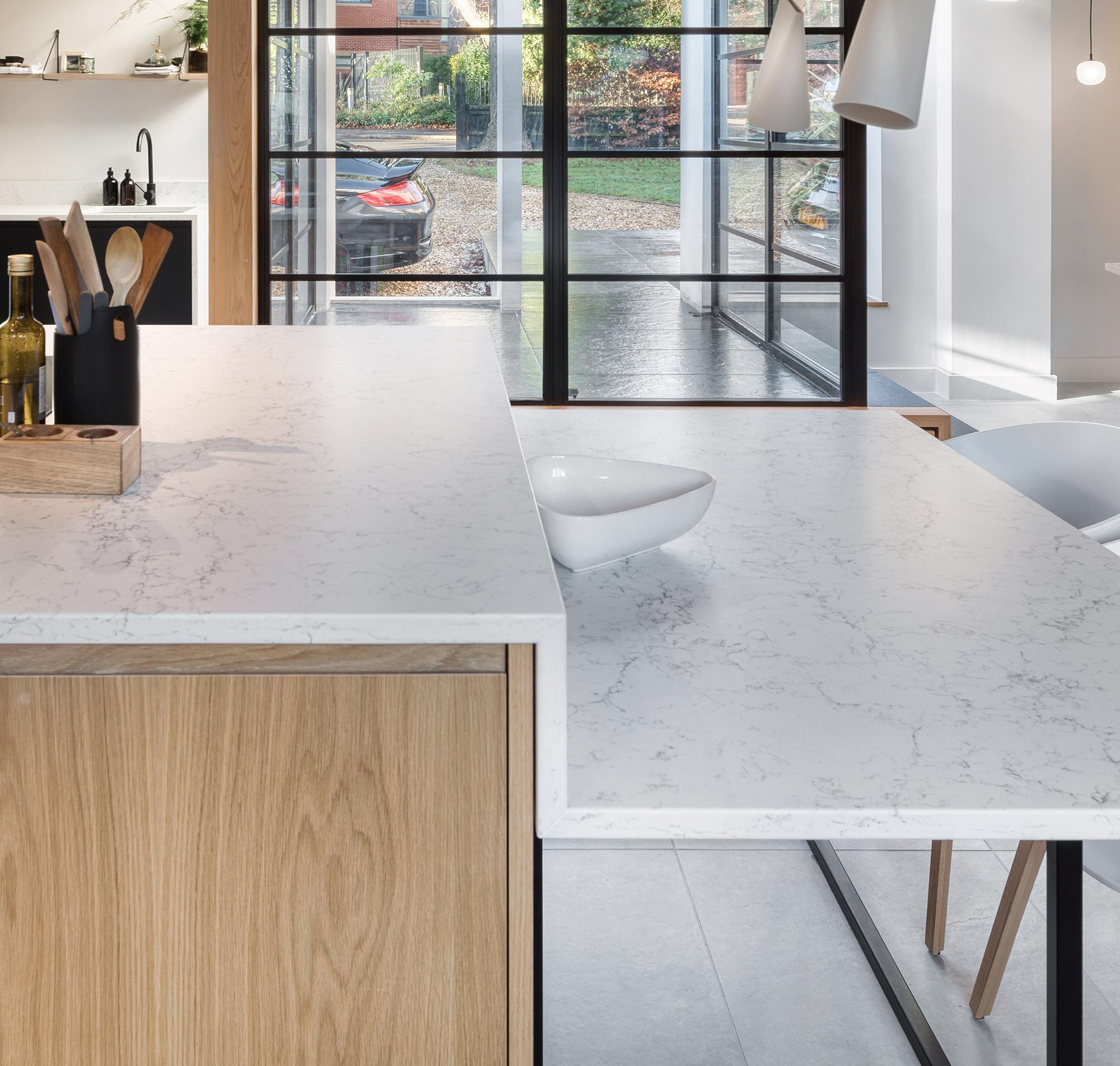 Modern kitchen with a white marble island, light wood cabinetry, a small white bowl on the counter, and large glass doors revealing a driveway and car outside. Sleek, minimalist decor with natural light.