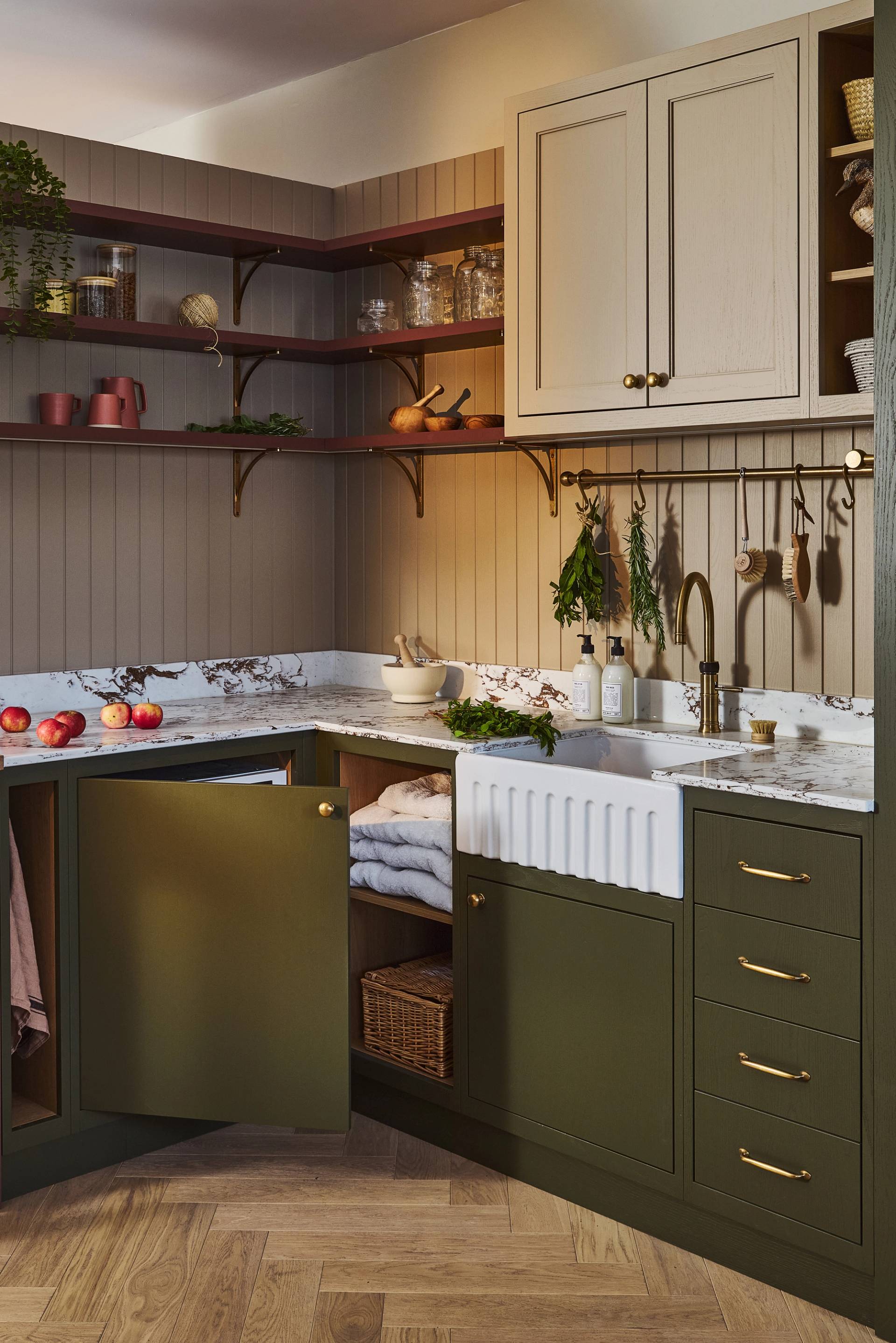 A stylish utility room with green cabinets, marble countertop, farmhouse sink, open shelves with dishes and plants, and neatly folded towels on a shelf.