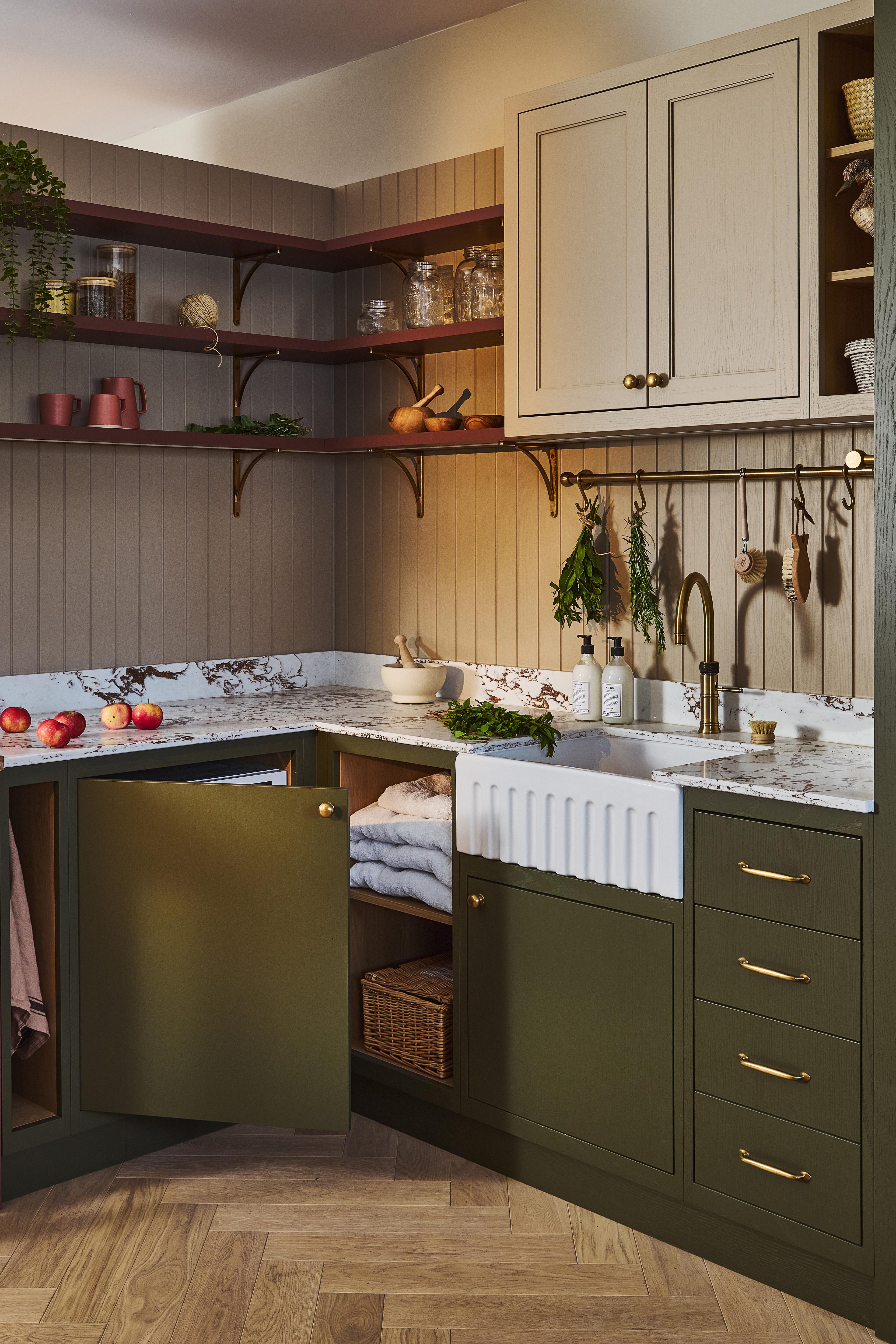 A stylish utility room with green cabinets, marble countertop, farmhouse sink, open shelves with dishes and plants, and neatly folded towels on a shelf.