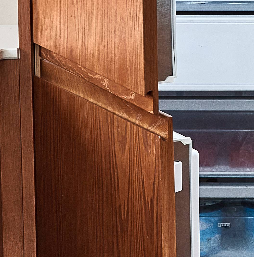 A wooden kitchen cabinet with two doors, one slightly open, revealing part of a white refrigerator with a visible lower freezer section. The cabinet and fridge are closely positioned together, suggesting a kitchen setting.