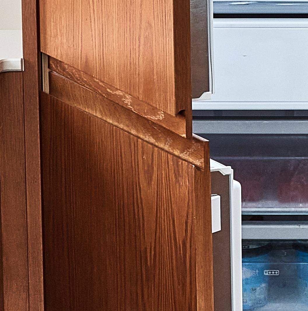 A wooden kitchen cabinet with two doors, one slightly open, revealing part of a white refrigerator with a visible lower freezer section. The cabinet and fridge are closely positioned together, suggesting a kitchen setting.
