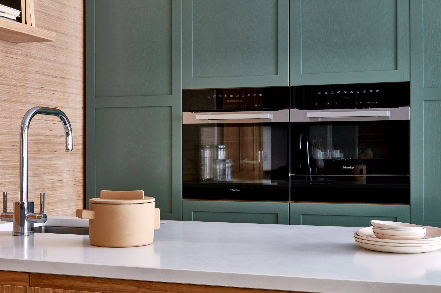 A modern kitchen with green cabinets, black built-in double ovens, and a beige ceramic pot with a lid on a white countertop. There is a stainless steel sink and a stack of beige plates next to the pot. The wall on the left has wooden paneling.