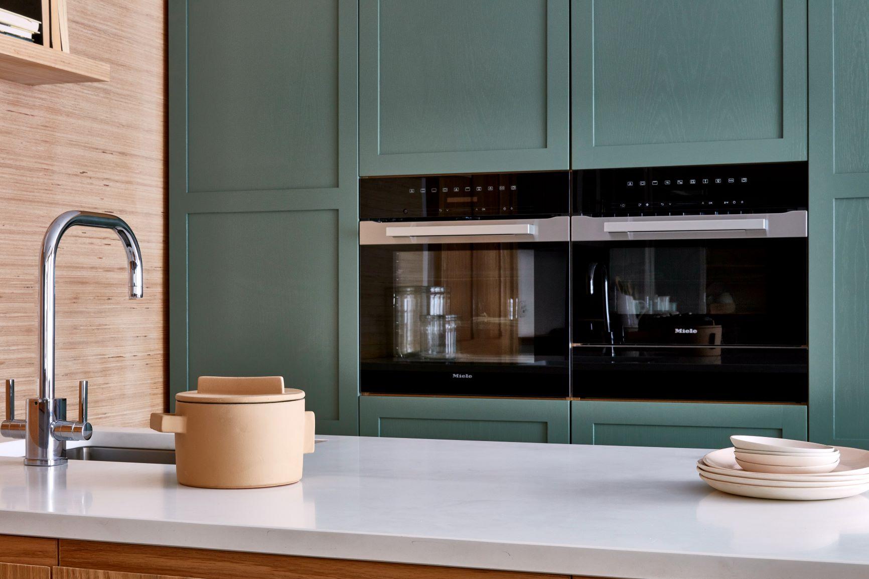A modern kitchen with green cabinets, black built-in double ovens, and a beige ceramic pot with a lid on a white countertop. There is a stainless steel sink and a stack of beige plates next to the pot. The wall on the left has wooden paneling.