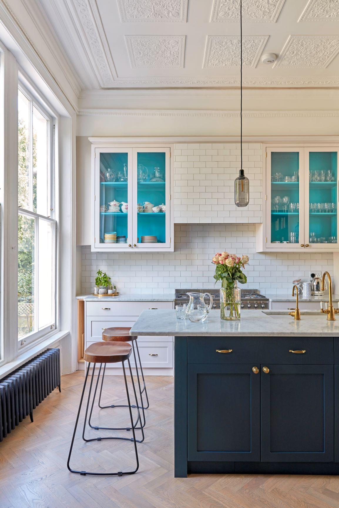 A bright kitchen with a large window, blue cabinets with glass doors, and a marble-topped island. Two wooden bar stools sit by the island, and a vase of pink roses is on the counter. A pendant light hangs above.