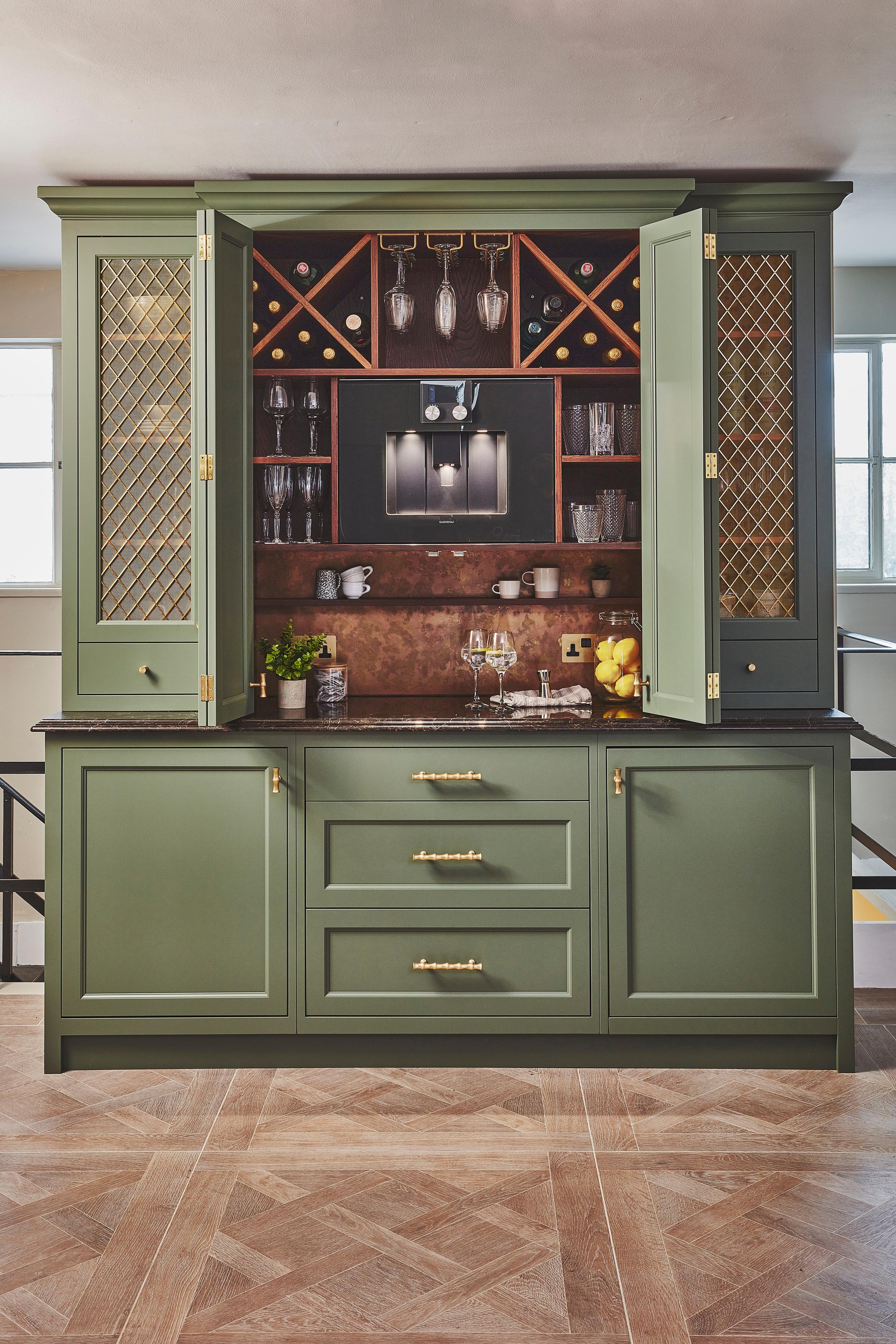 Green kitchen cabinet with gold handles and mesh-paneled doors, open to reveal a built-in espresso machine, wine storage, glassware, and shelves holding cups and bottles, set against a brass backsplash.