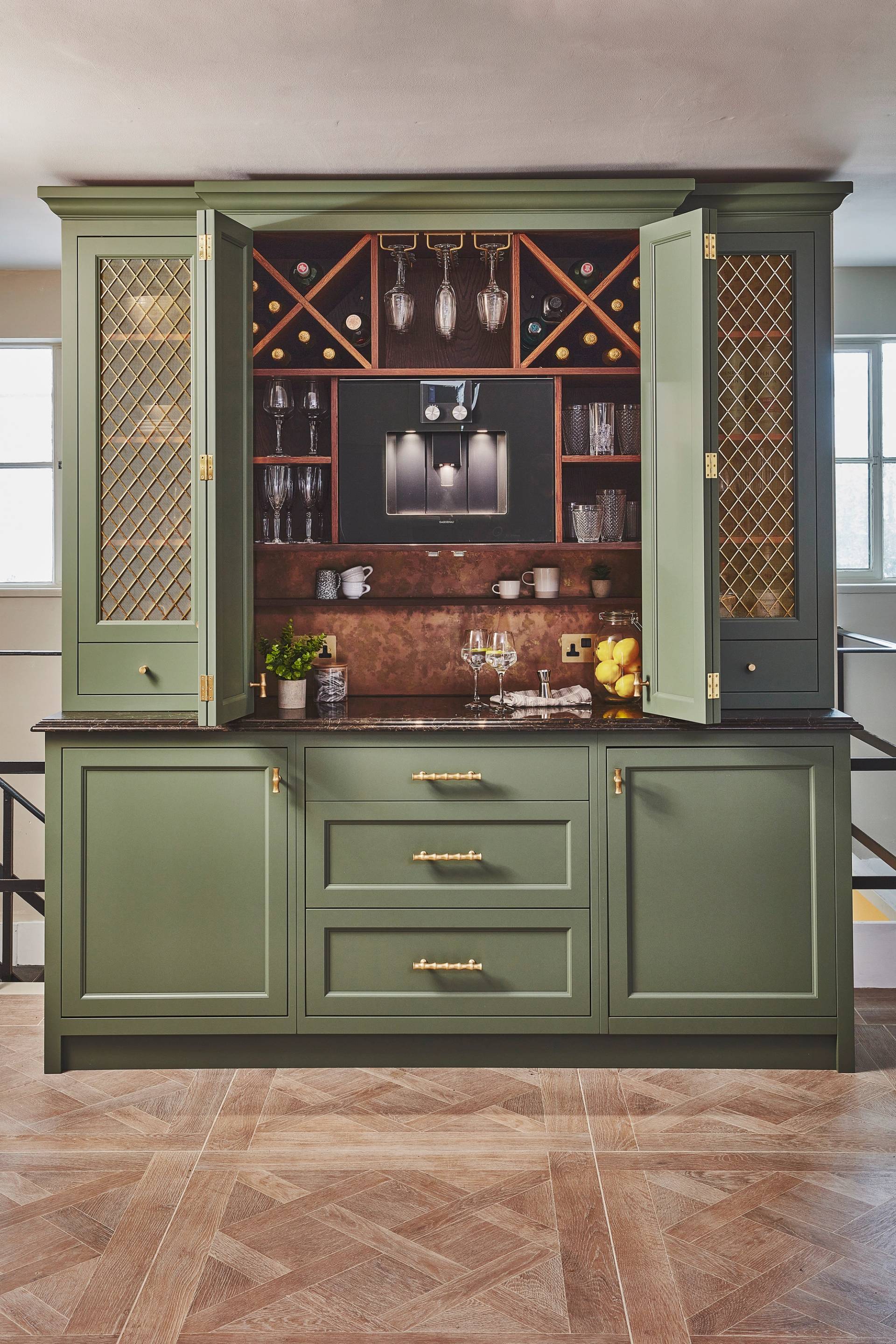 Green kitchen cabinet with gold handles and mesh-paneled doors, open to reveal a built-in espresso machine, wine storage, glassware, and shelves holding cups and bottles, set against a brass backsplash.