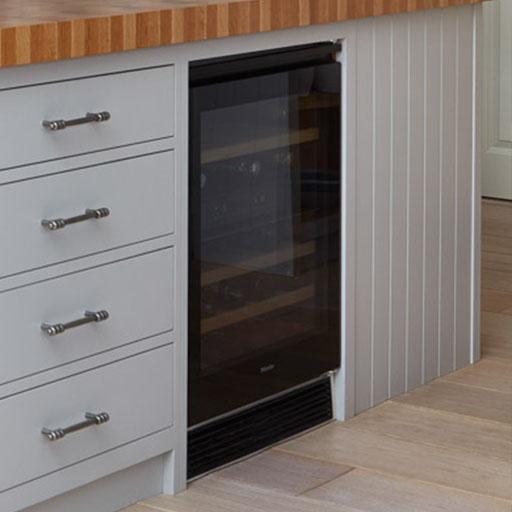 A built-in wine cooler with a glass door is installed under a wooden countertop next to gray drawers with silver handles in a modern kitchen.