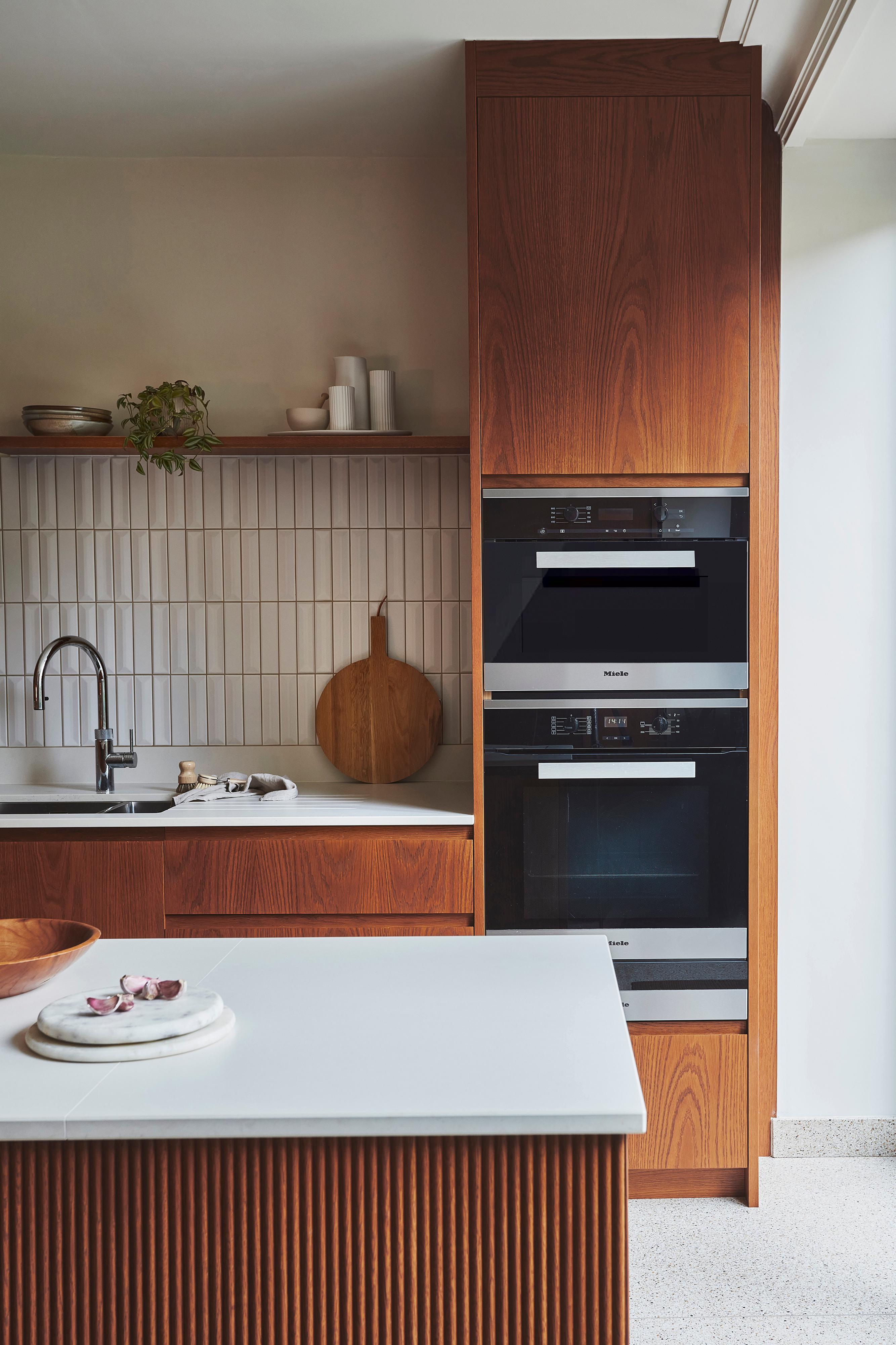 Modern kitchen with sleek wood cabinetry, a wall-mounted oven, and a microwave. The backsplash features vertical white tiles, and a wooden cutting board rests on the counter. A small plant and decorative items sit on a shelf.