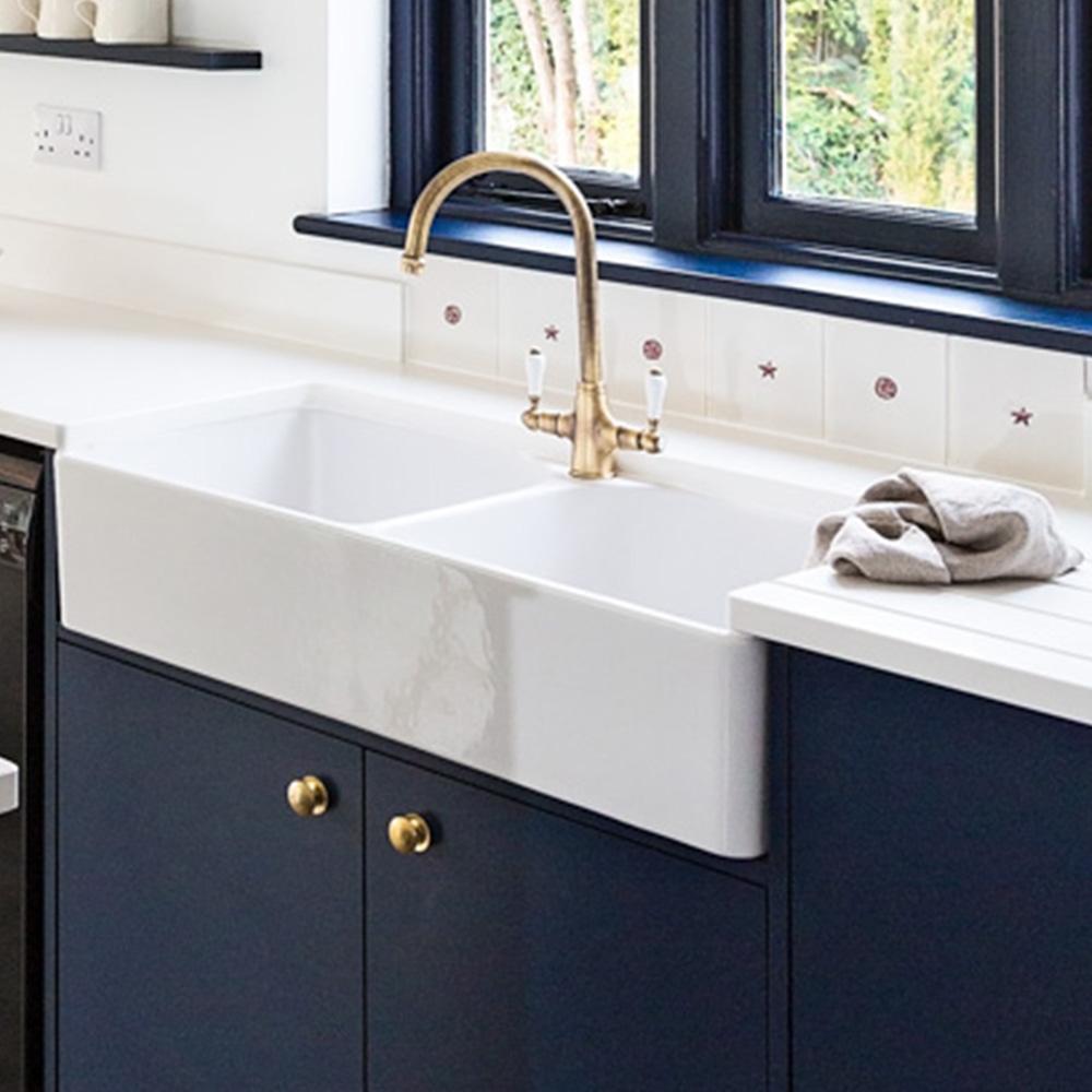 A modern kitchen with a white farmhouse sink, brass faucet, dark blue cabinets with brass knobs, white countertop, and a towel on the side. A window with black trim overlooks greenery outside.