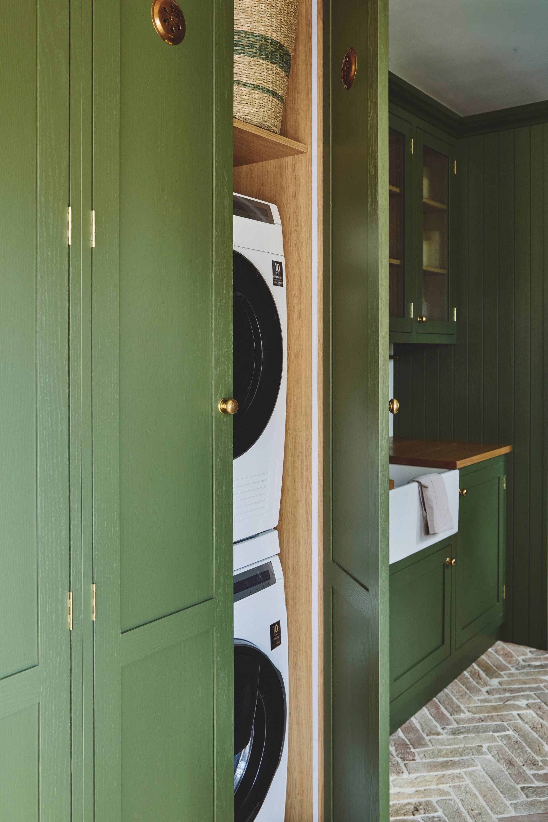 A laundry area with olive green cabinets, a stacked washer and dryer inside a closet, woven basket above, a farmhouse sink, and herringbone patterned tile floor.