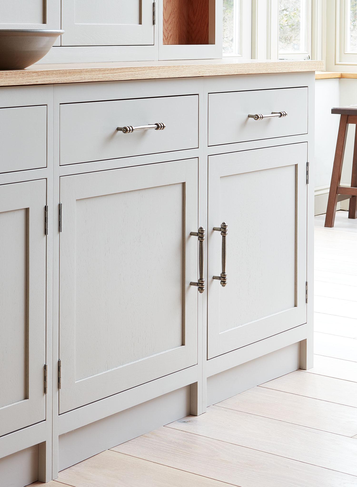 A close-up of light gray kitchen cabinets with silver handles and a light wood countertop, set in a bright room with pale wood flooring and large windows in the background.