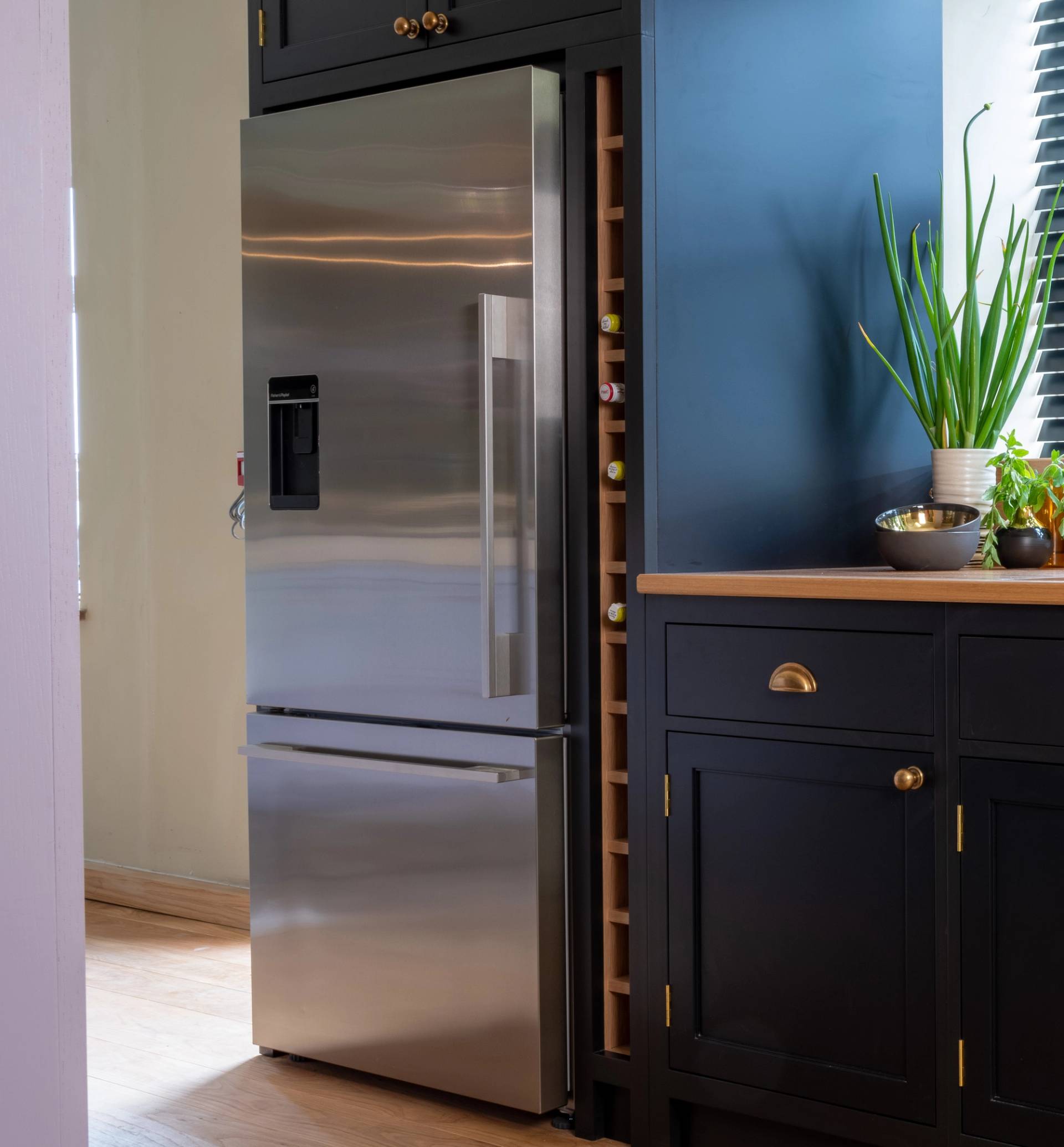 A modern stainless steel refrigerator with a built-in water dispenser stands beside dark cabinets. A small wine rack is integrated on the side, and a plant and bowl decorate the wooden countertop. Sunlight streams in through nearby blinds