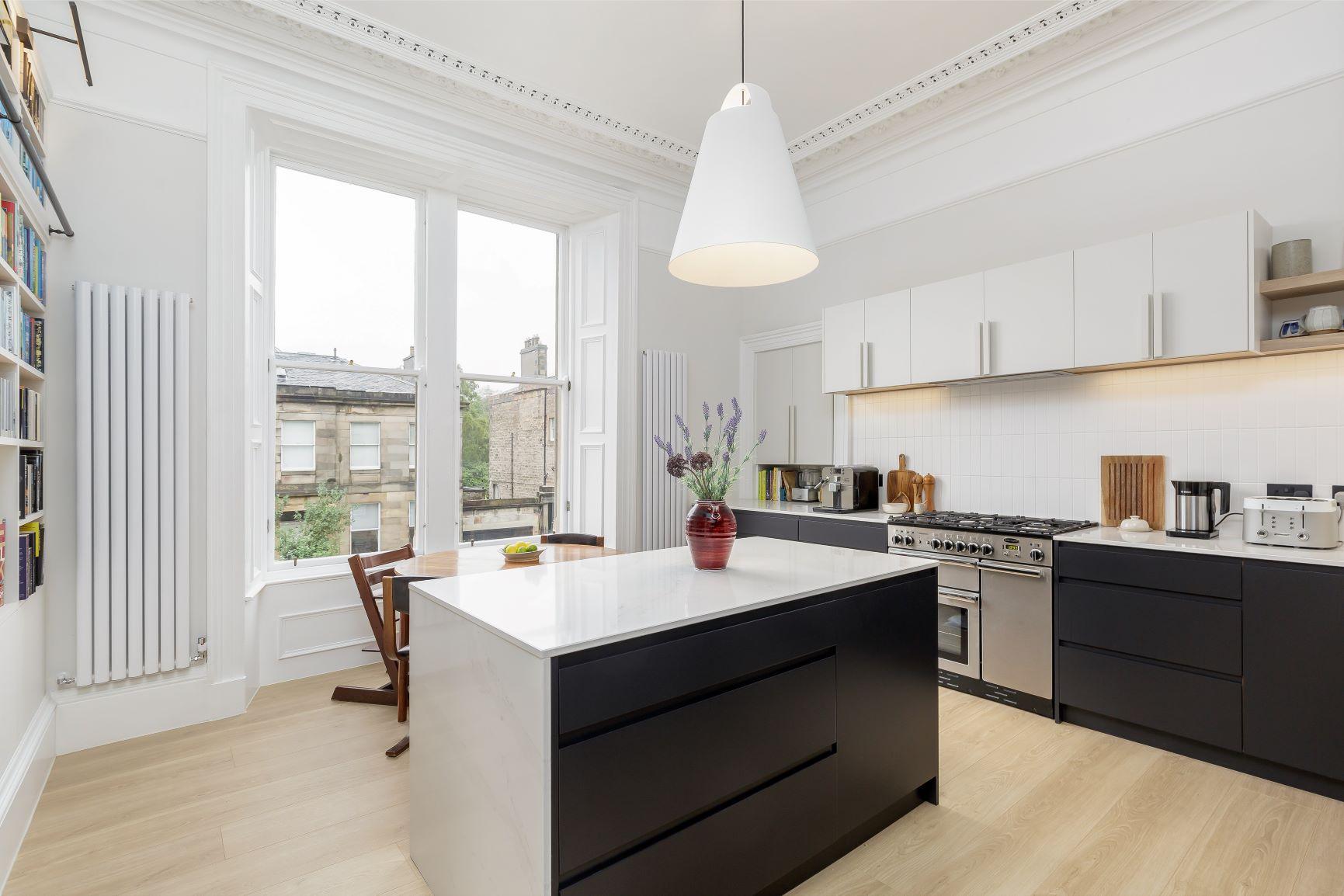 A modern kitchen with white walls, a large window, and light wood floors. It features a black and white island with a potted plant, sleek cabinets, a stainless steel stove, and contemporary lighting. A chair sits by the window.