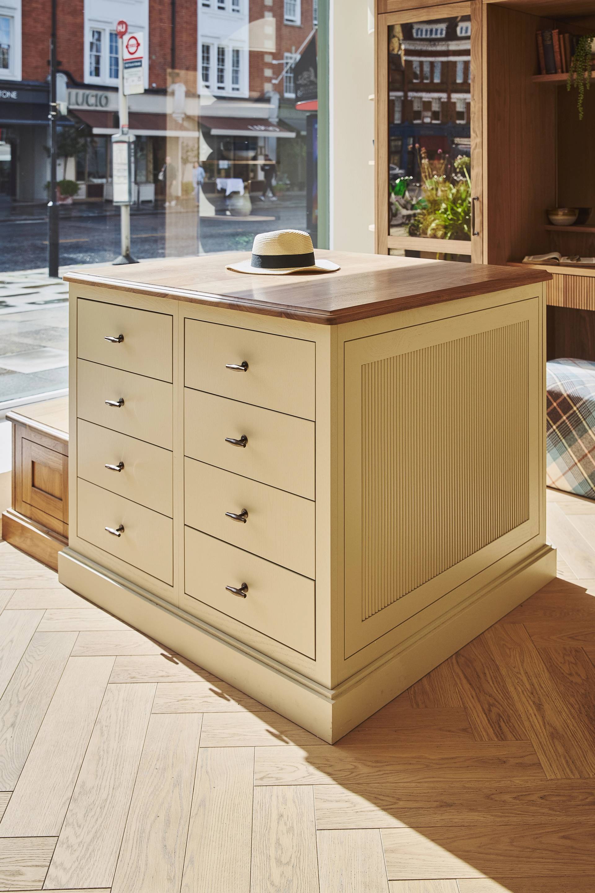 A beige wooden cabinet with multiple drawers stands by a window in a sunlit room. A hat rests on its wooden countertop. The background shows a street scene and shops outside.