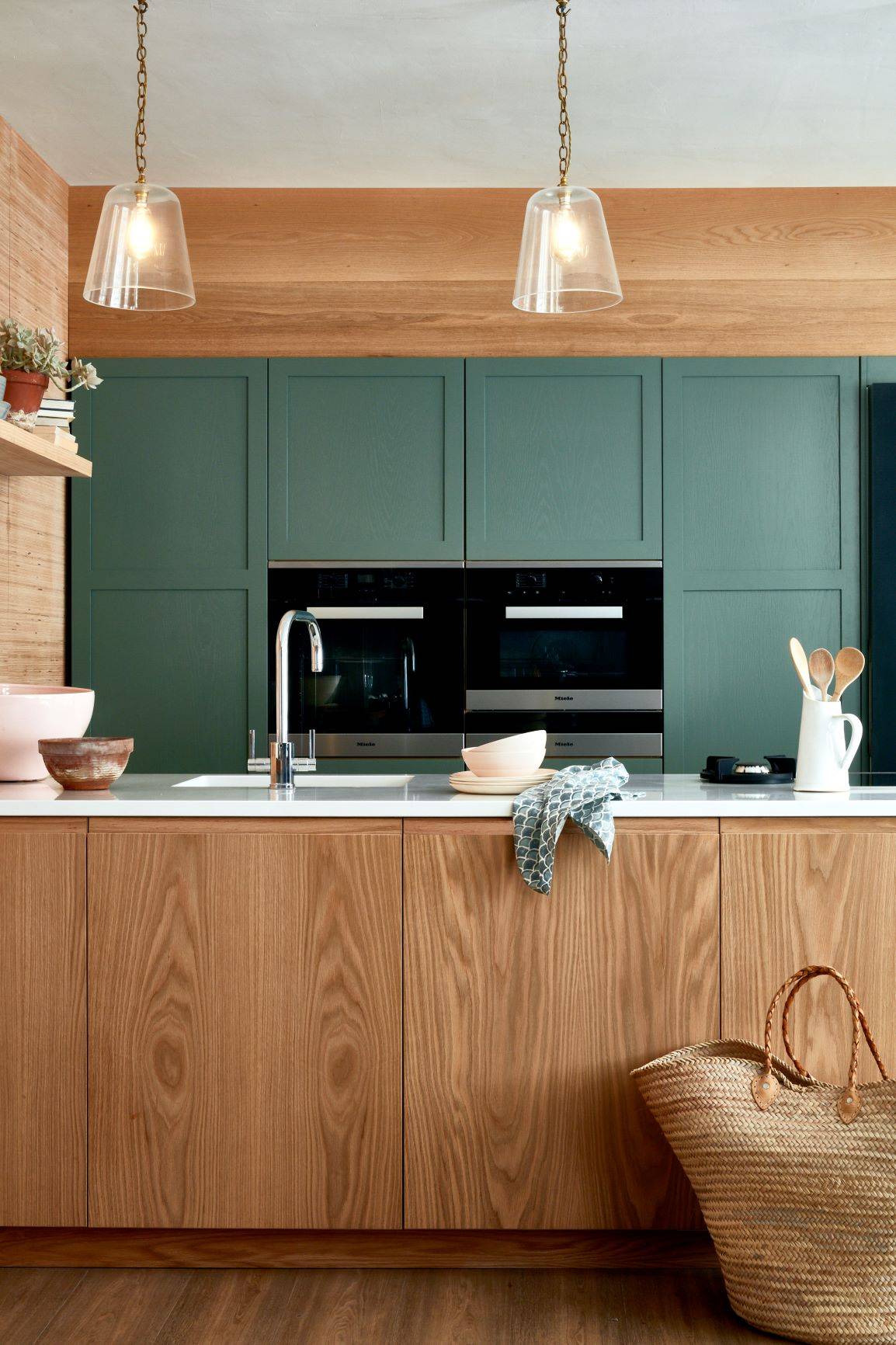 A modern kitchen with wood grain cabinets and a green accent wall. Hanging glass pendant lights illuminate the space. A woven basket sits on the floor, and various kitchen items are arranged on the counter.