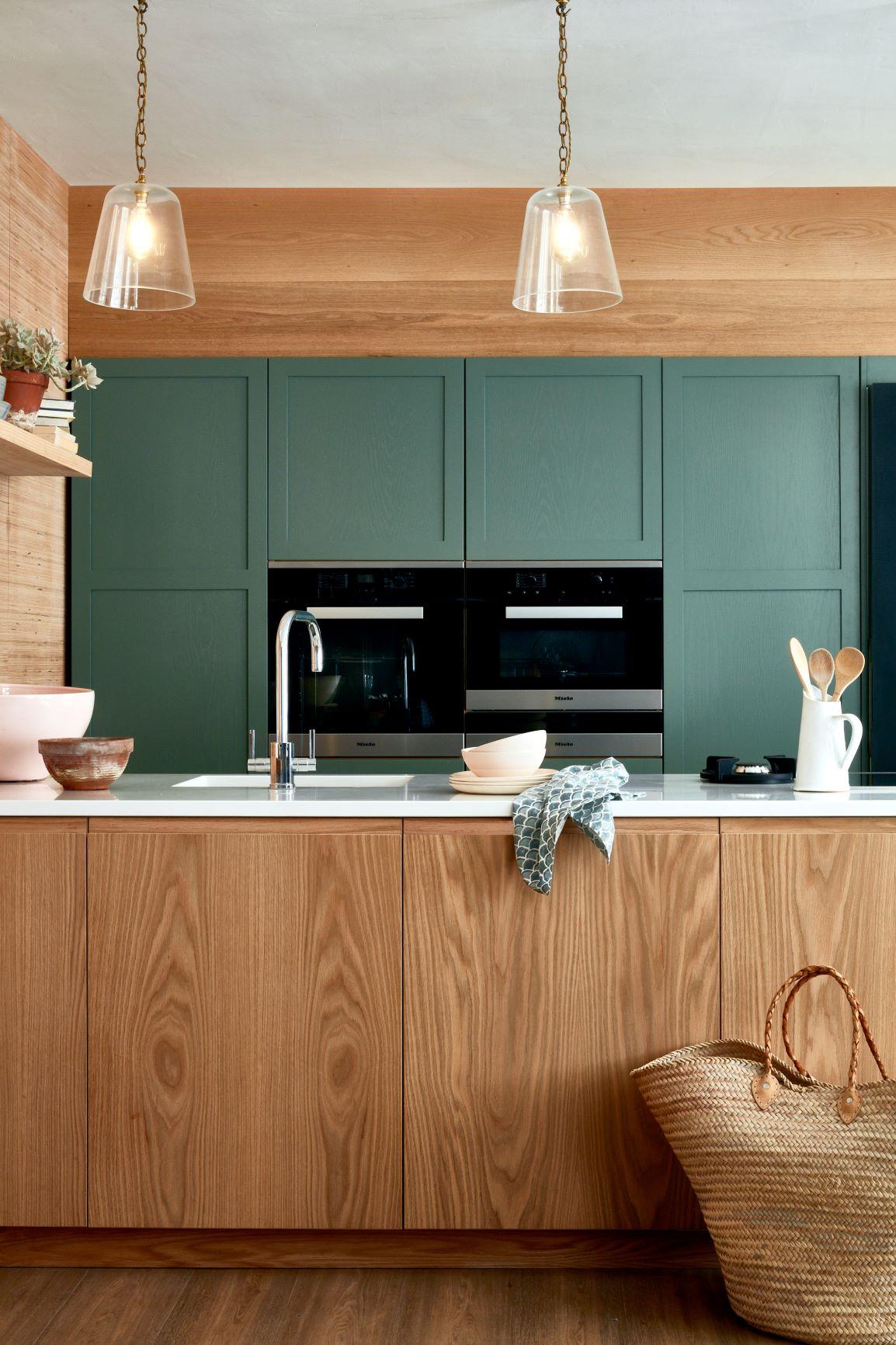 A modern kitchen with wood grain cabinets and a green accent wall. Hanging glass pendant lights illuminate the space. A woven basket sits on the floor, and various kitchen items are arranged on the counter.