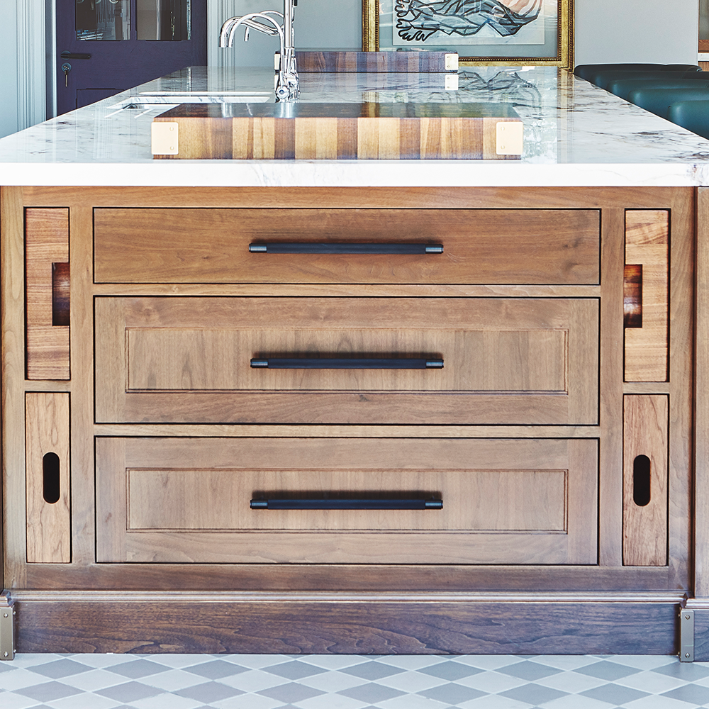 A wooden kitchen island with three large drawers featuring black handles, a marble countertop with a built-in sink, and patterned tile flooring.
