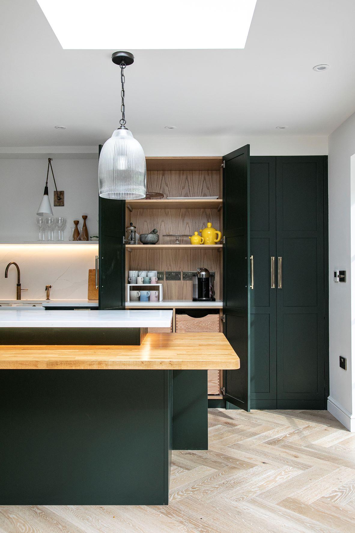 Modern kitchen with dark green cabinets, wooden countertops, open cabinet displaying dishware and yellow teapots, herringbone wood floors, and a pendant light hanging from the ceiling.
