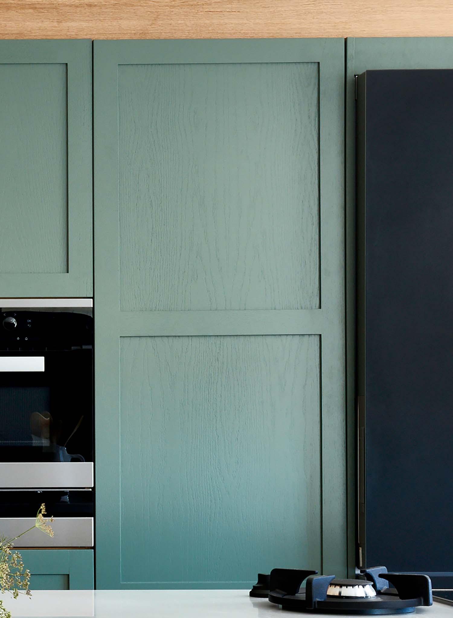 A close-up view of a modern kitchen featuring matte green cabinet doors with a subtle wood grain texture, a built-in stainless steel oven, and a black gas stove on a white countertop.