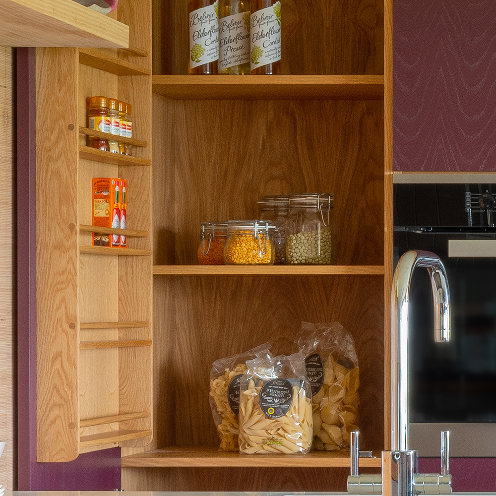 An open wooden kitchen cupboard displays glass jars with grains, bags of pasta, spice jars on a rack, and bottles on the top shelf. A metal sink faucet is visible in the foreground.