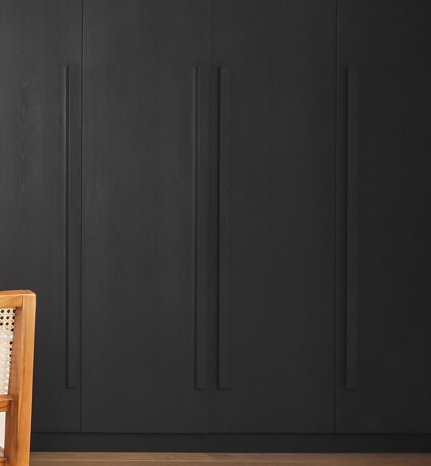 A partial view of a wooden chair with a woven seat and backrest, positioned in front of large, matte black cabinets with long vertical handles. The floor is brown wood.