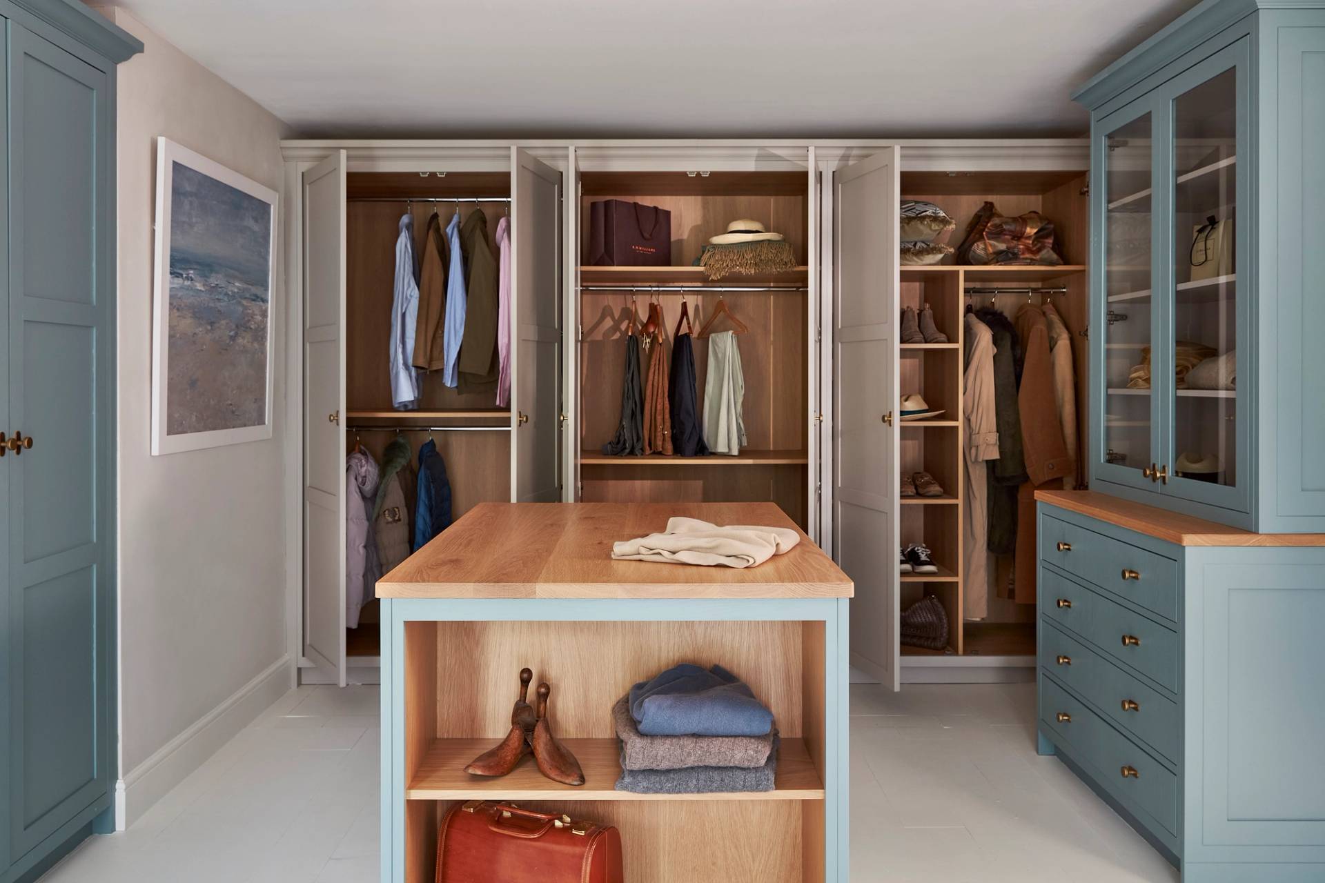 A spacious, organized walk-in closet with open cabinets showing hanging clothes, shelves with shoes, bags, and hats, and a wooden island in the center holding folded clothes and a pair of boots.