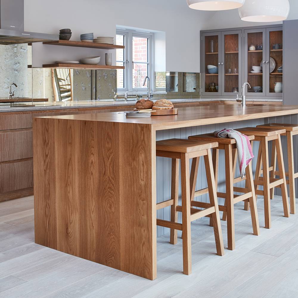 Modern kitchen with a large wooden island, four wooden bar stools, loaves of bread on the countertop, mirrored backsplash, open shelving, and cabinets with glass doors. Light streams in from a window.