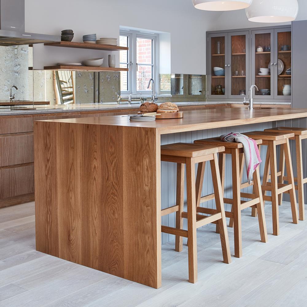 Modern kitchen with a large wooden island, four wooden bar stools, loaves of bread on the countertop, mirrored backsplash, open shelving, and cabinets with glass doors. Light streams in from a window.