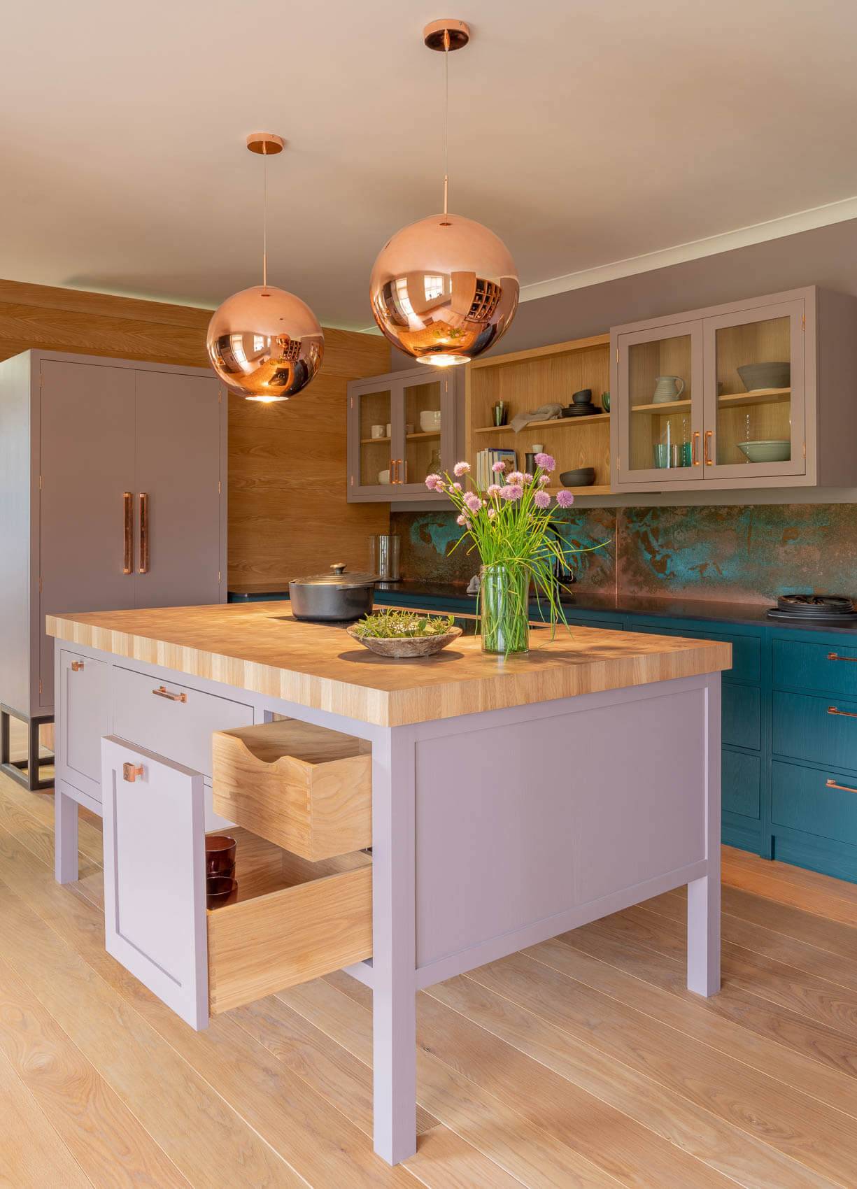 A modern kitchen with a wooden island features open drawers and vases of flowers. Copper pendant lights hang above. The kitchen has a combination of grey and teal cabinets, with glass-fronted cupboards and a metallic backsplash.