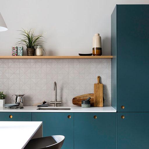 Modern kitchen with teal cabinets, white countertops, and a geometric-patterned backsplash. A wooden shelf above the sink holds plants, cups, and jars; cutting boards and a teapot sit on the counter.
