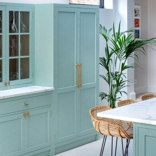 Light blue kitchen cabinets with gold handles, a white countertop, a tall green plant, and wicker bar stools at a marble-topped island in a bright, modern kitchen.