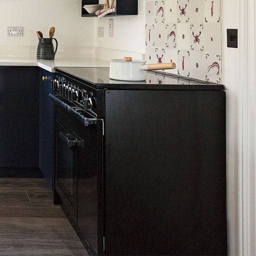 A black stove with an oven stands against a white wall, topped with a white pot and wooden spoon; the backsplash has a pattern of sea creatures, and the countertop holds a dark pitcher with utensils.