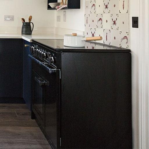 A black stove with an oven stands against a white wall, topped with a white pot and wooden spoon; the backsplash has a pattern of sea creatures, and the countertop holds a dark pitcher with utensils.