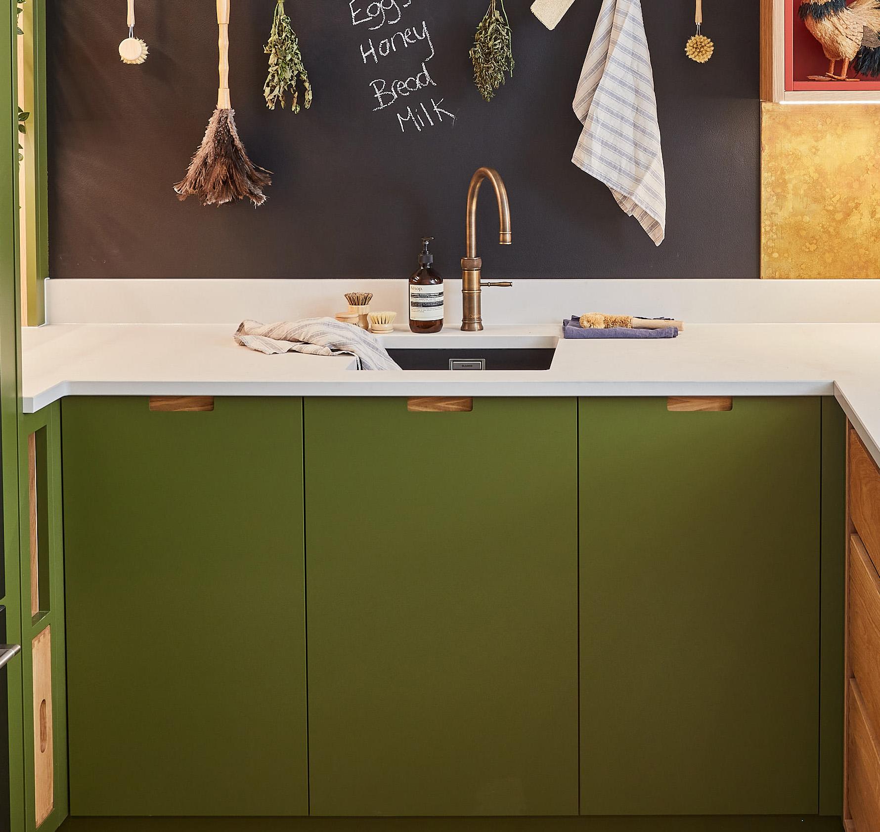 A kitchen corner with olive green cabinets and a white countertop. A black sink and bronze faucet are centered. Dried herbs and a towel hang above. A chalkboard wall lists Eggs, Honey, Bread, Milk. A soap dispenser and folded cloth are near the sink.