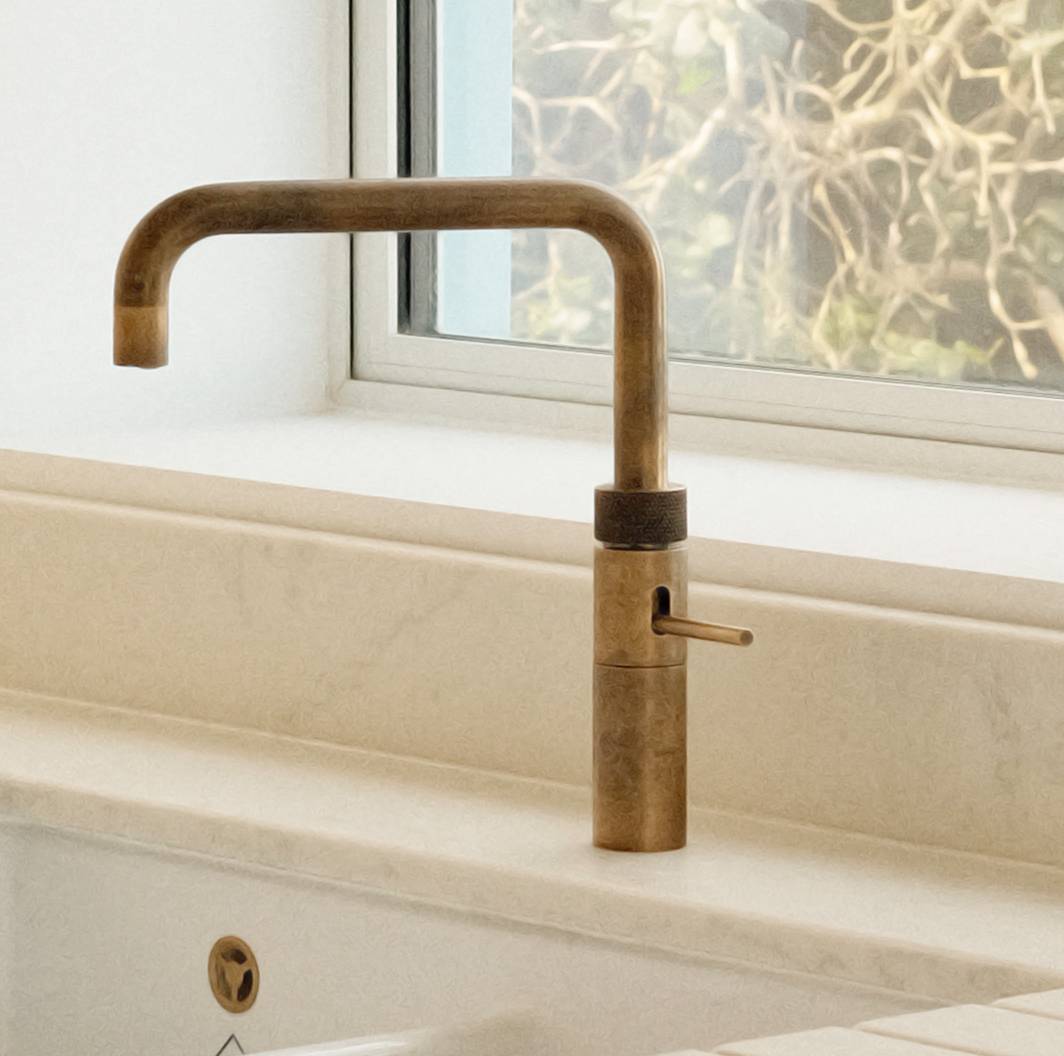 A vintage-style brass kitchen faucet with a right-angled spout is installed on a light-colored countertop near a window with a blurred outdoor view.