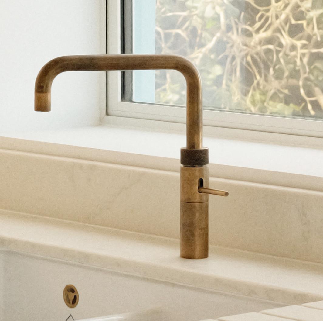 A vintage-style brass kitchen faucet with a right-angled spout is installed on a light-colored countertop near a window with a blurred outdoor view.