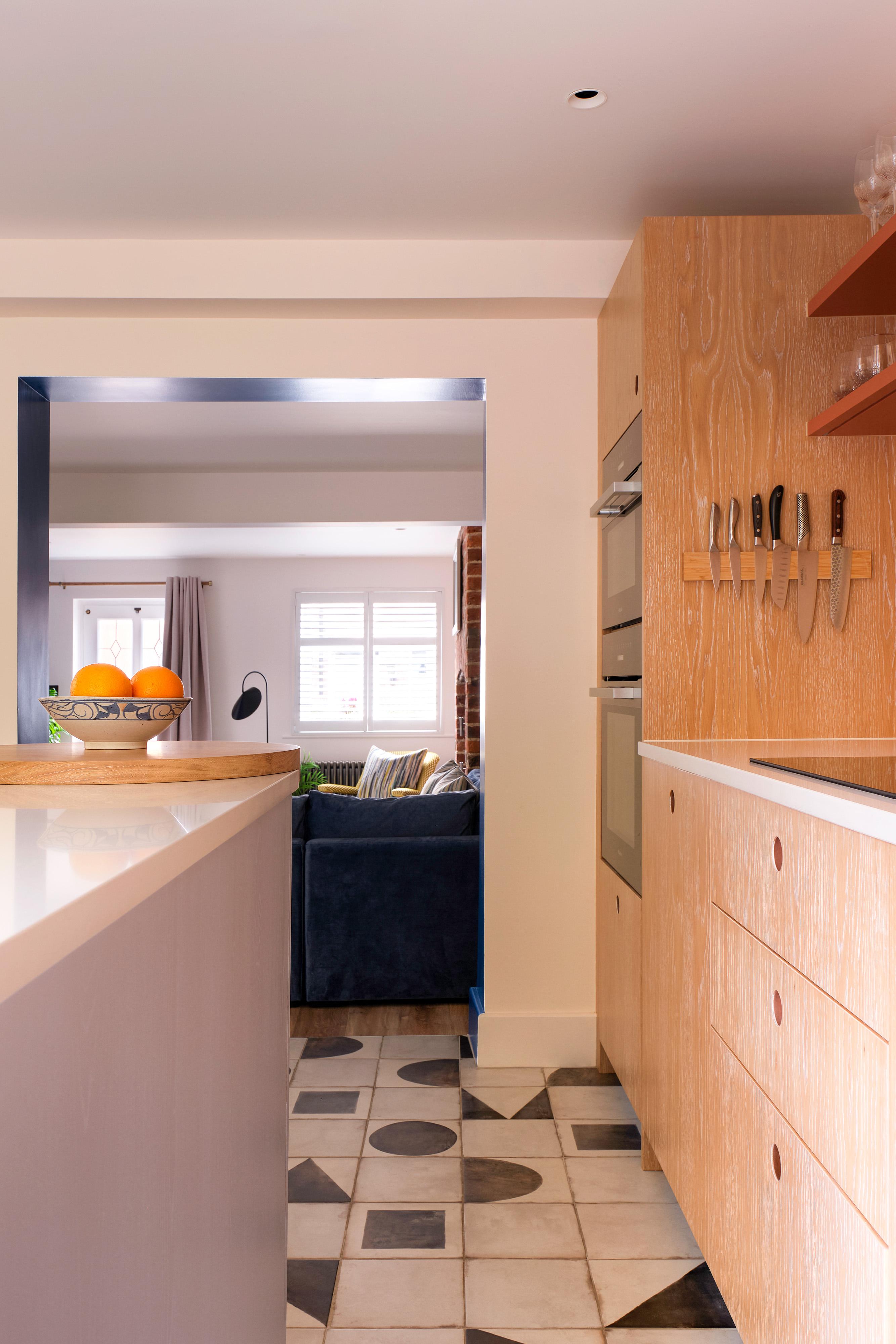 A modern kitchen with wooden cabinets, built-in oven, knives on a magnetic strip, and a bowl of fruit on the counter, leading into a living room with a blue sofa and window