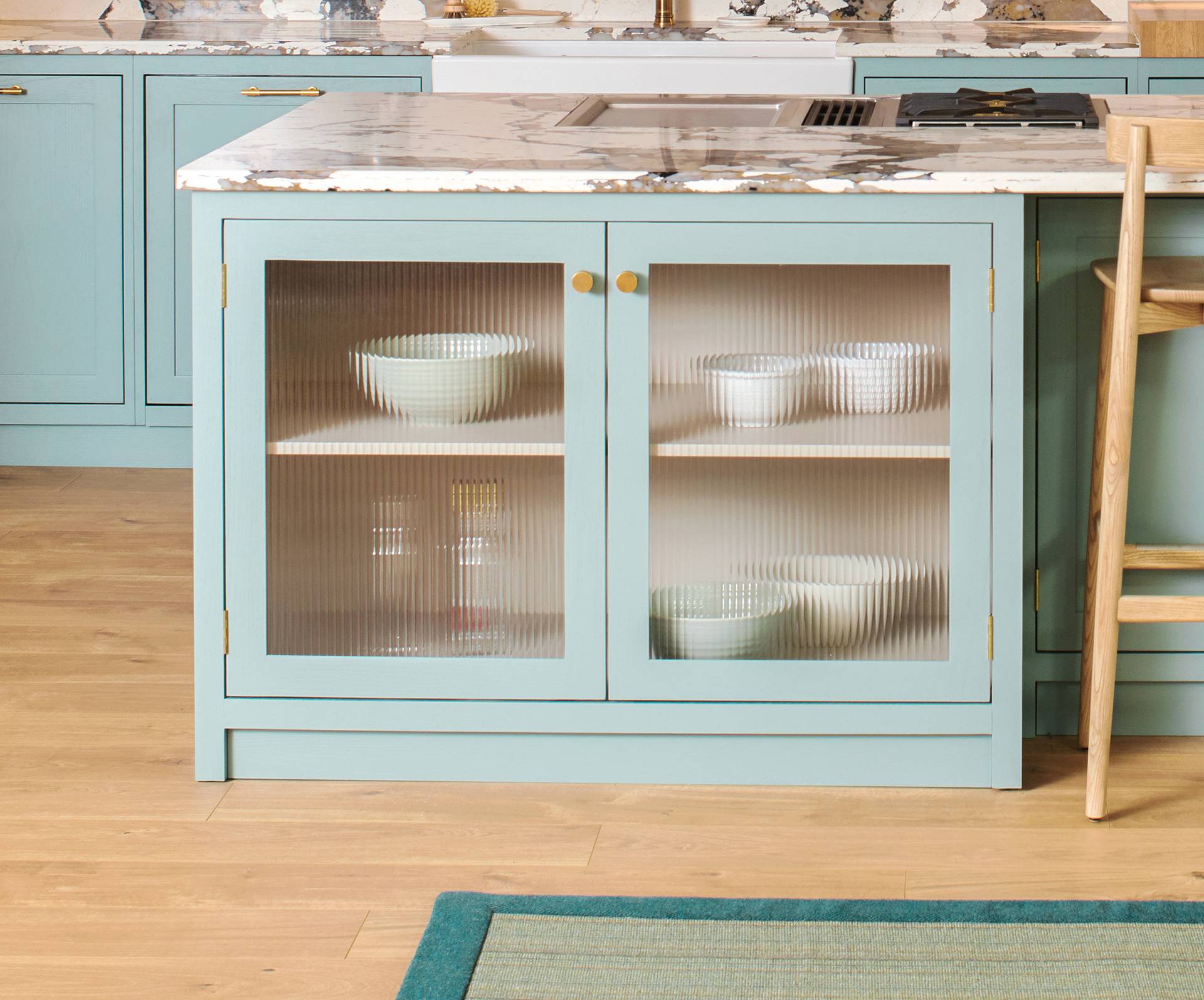 A kitchen island with light blue cabinetry featuring frosted glass doors. Inside, there are white bowls on wooden shelves. The countertop is marble, and the floor is light wood. Part of a barstool and a green rug is visible.