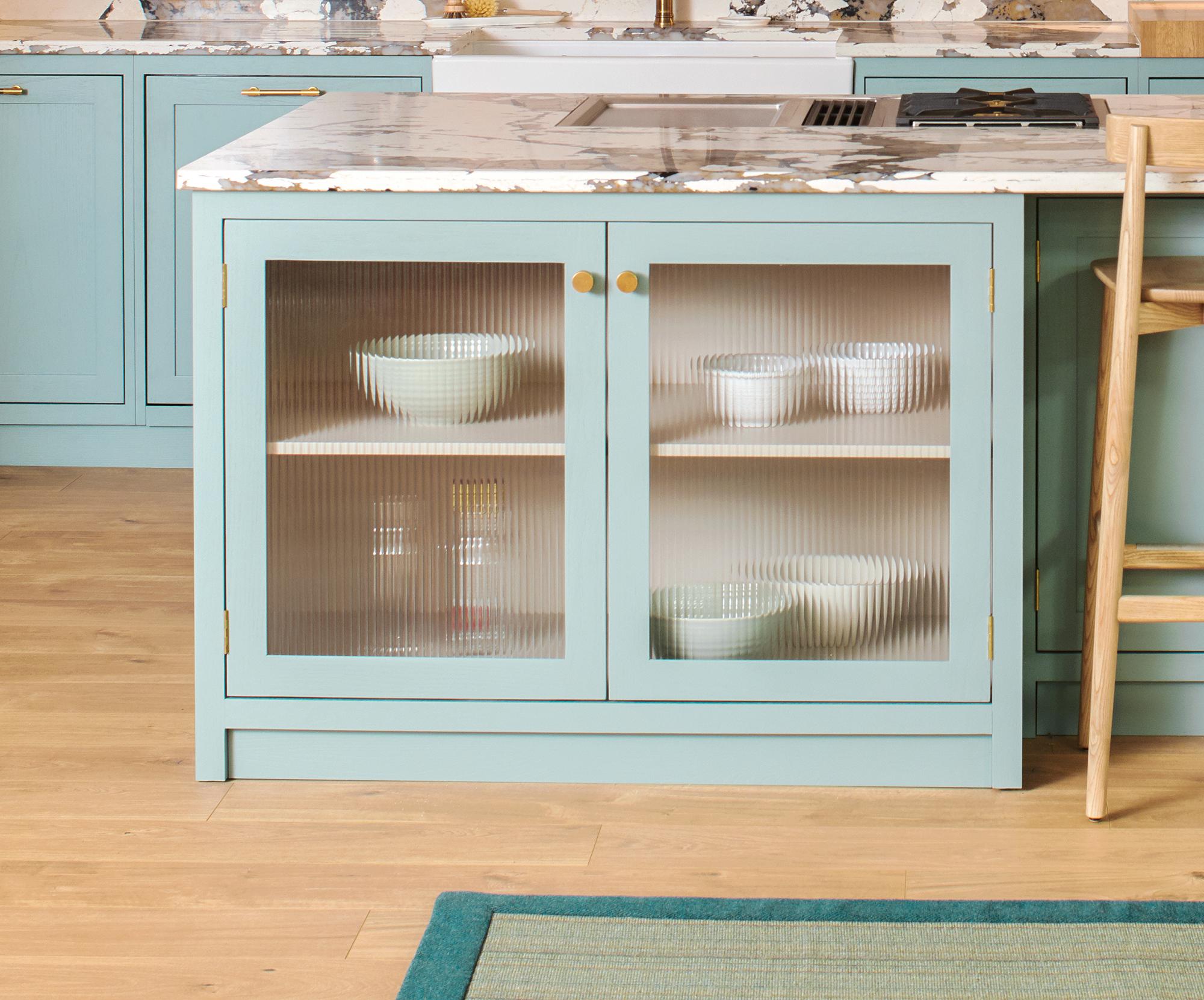 A kitchen island with light blue cabinetry featuring frosted glass doors. Inside, there are white bowls on wooden shelves. The countertop is marble, and the floor is light wood. Part of a barstool and a green rug is visible.